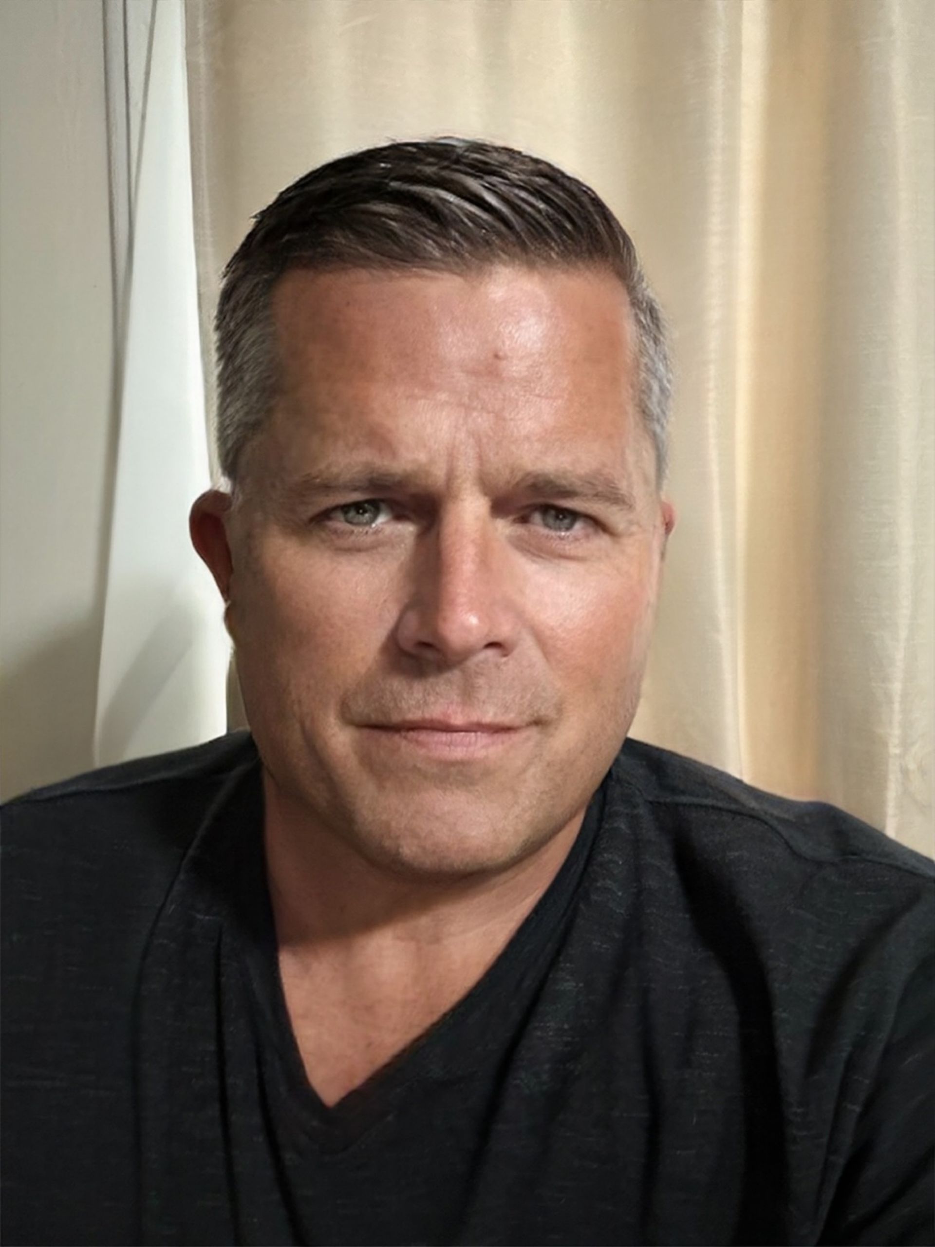 Man in a black shirt with a slight smile, seated indoors in front of beige curtains.