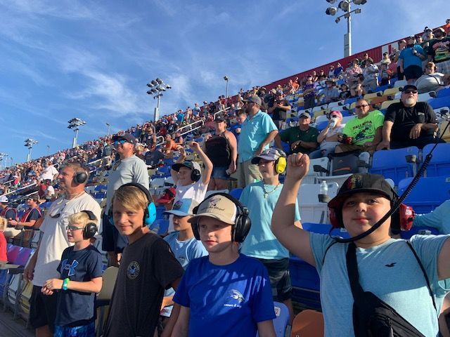 Crowd of fans in blue stadium seats, some standing and cheering under a clear sky