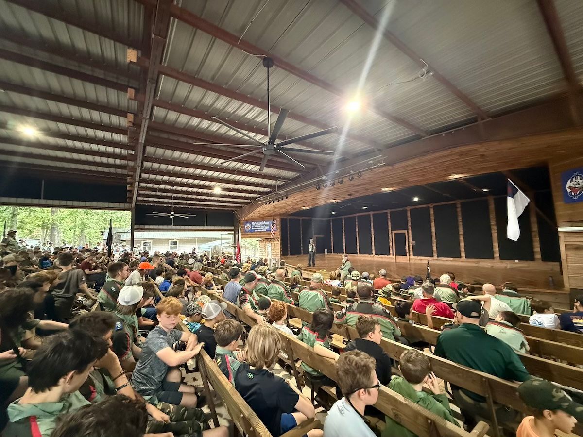 Crowded covered grandstand with people seated under a metal roof, facing a stage or arena area