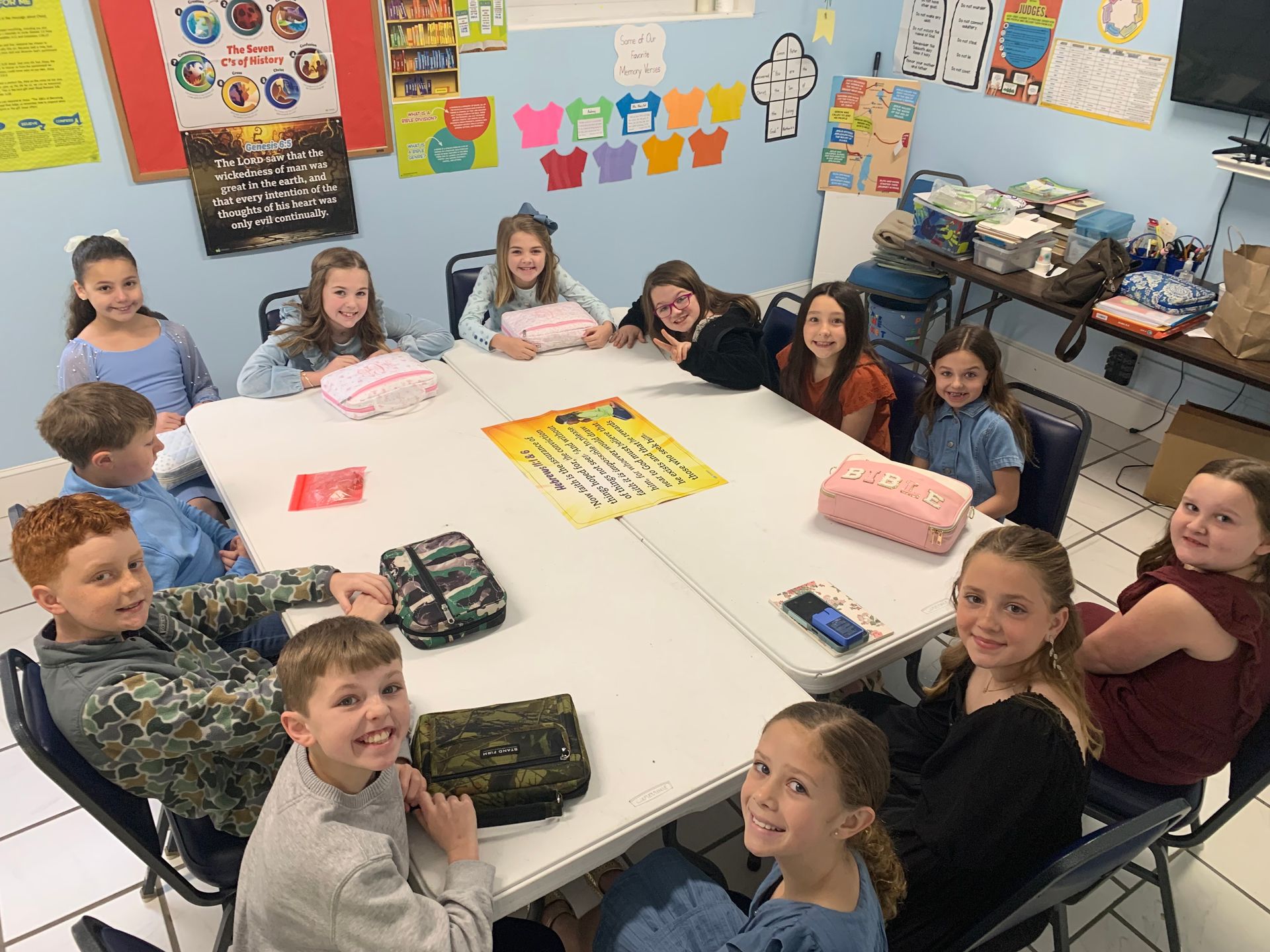 Students sit around a white table in a colorful classroom, with gifts and cards on the table.