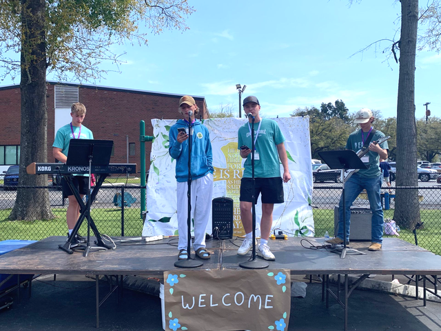 Four musicians performing on an outdoor stage with microphones and instruments, one playing keyboard.