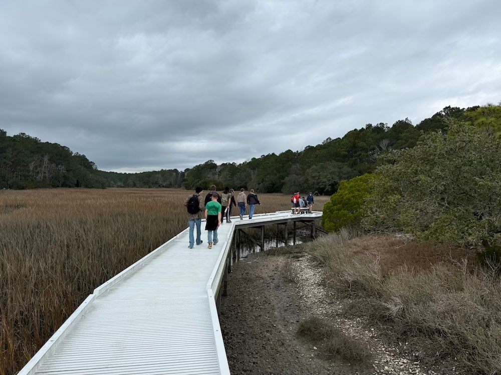 People walking on a white boardwalk through a marsh under a cloudy sky