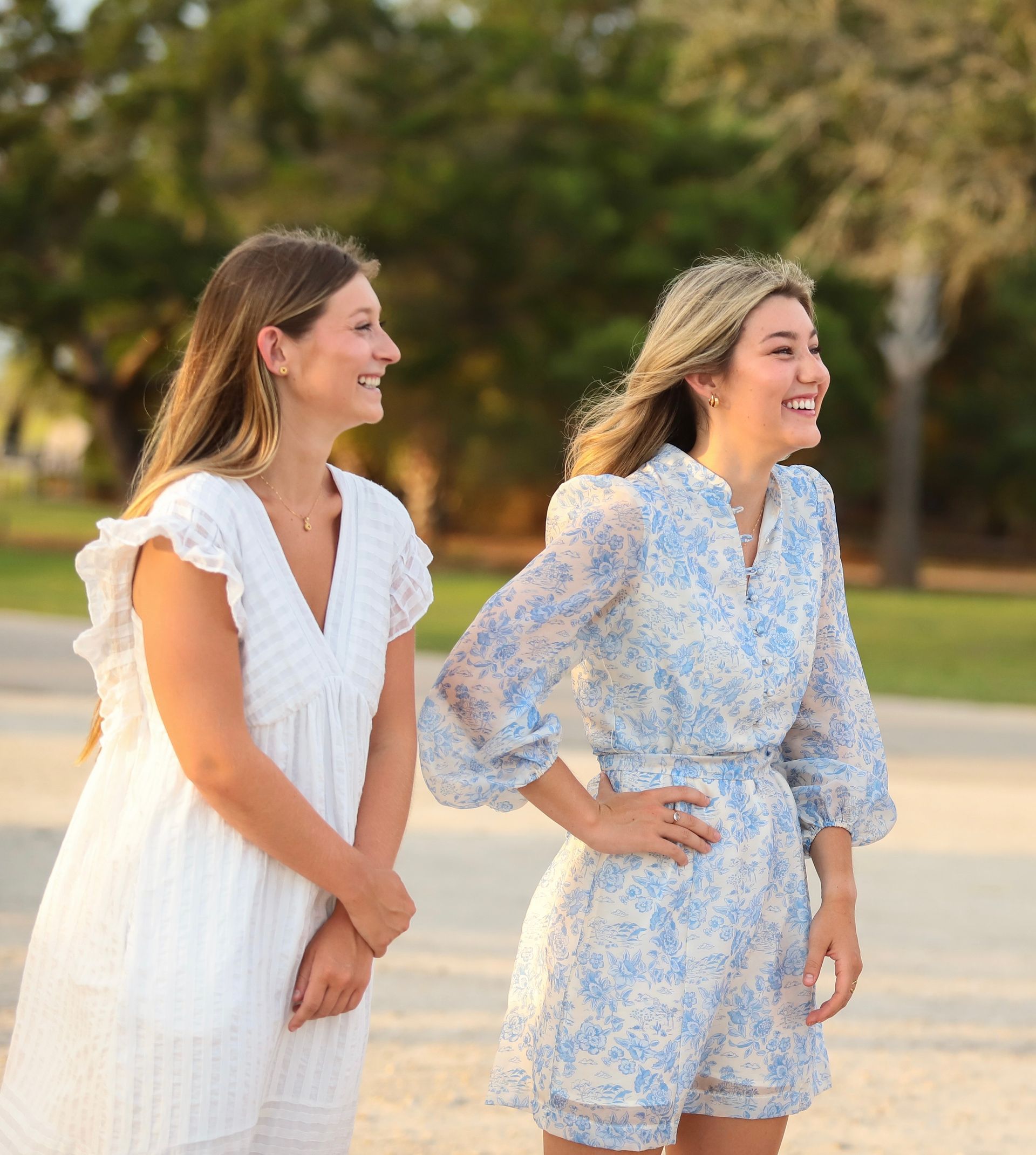 Two women smiling outdoors, one in white and one in a blue floral dress, standing in a park