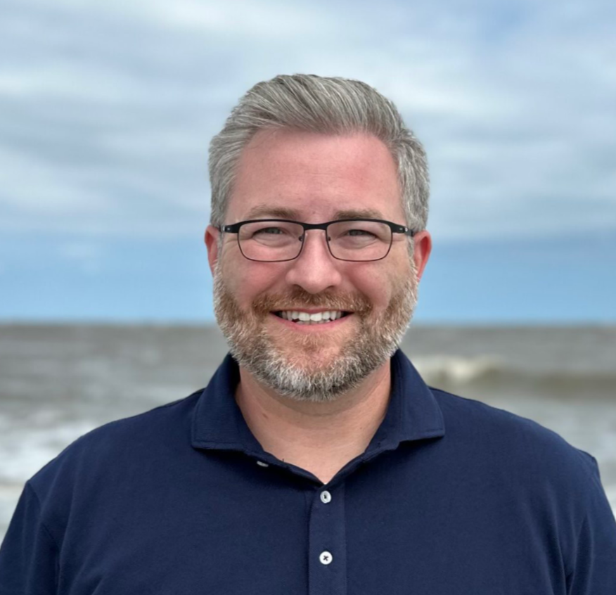 Smiling man in glasses and navy polo standing outdoors near the ocean