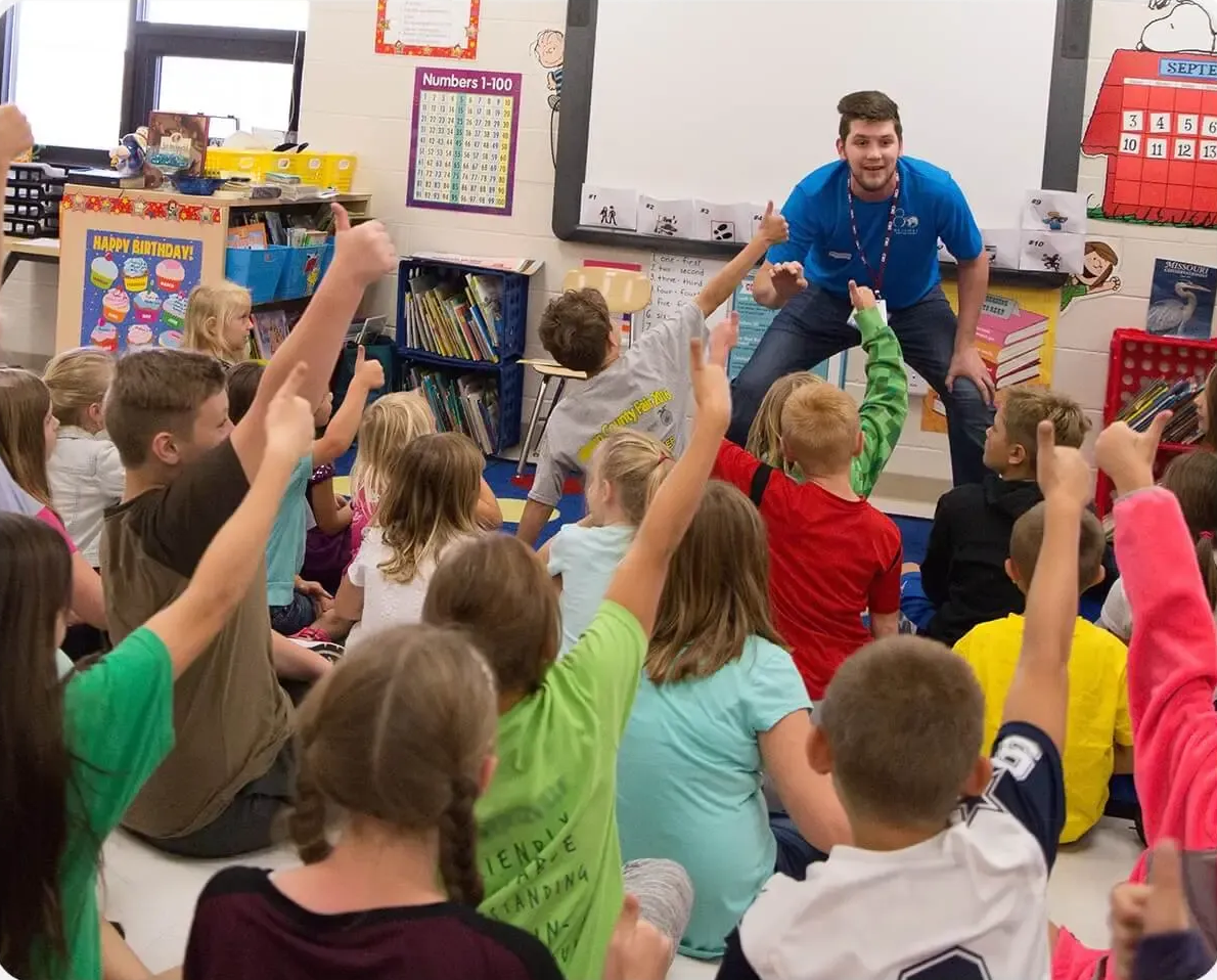 Teacher speaking to seated children in a classroom, with several students raising their hands.