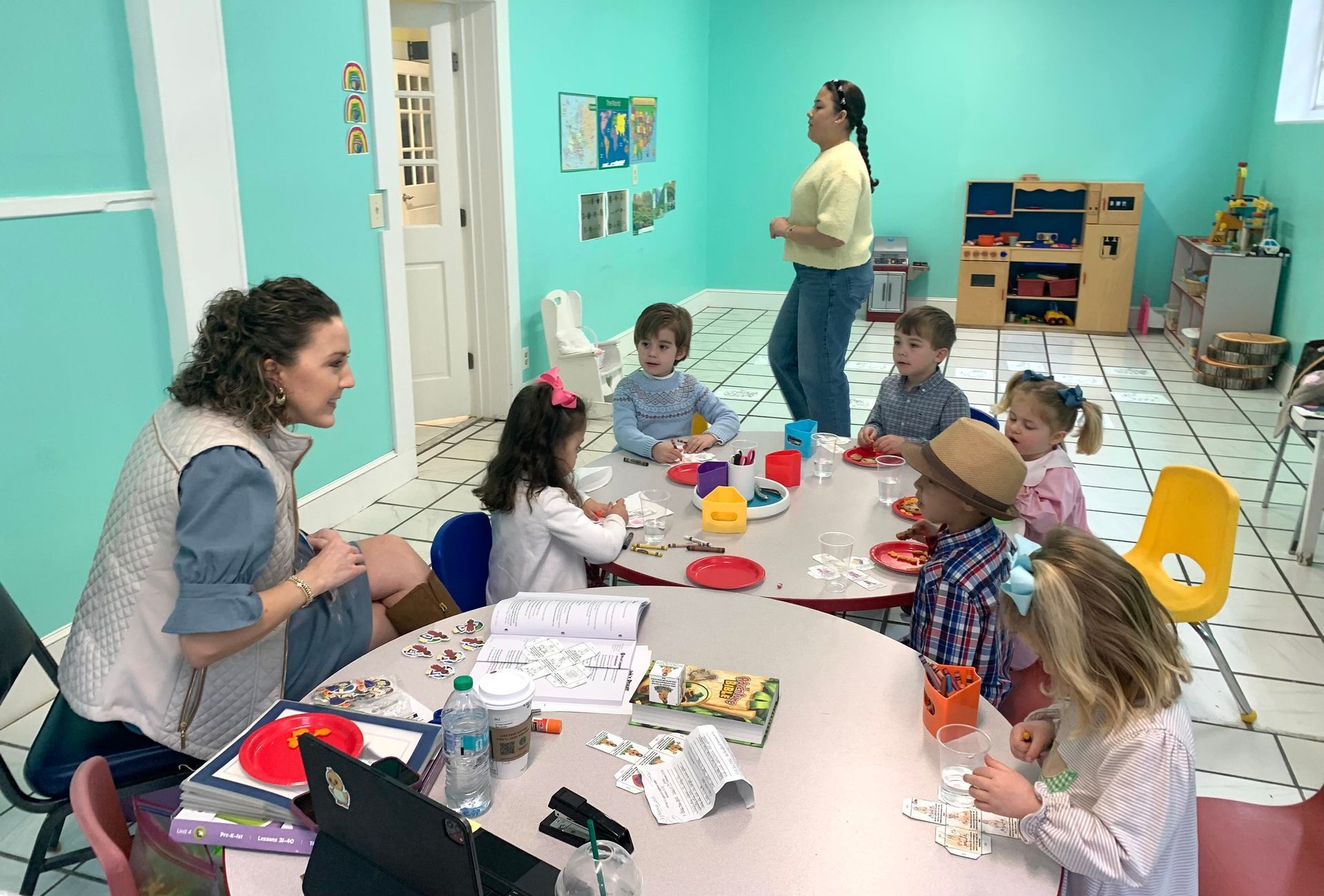Children and adults gathered around a table in a bright classroom, making a craft activity.