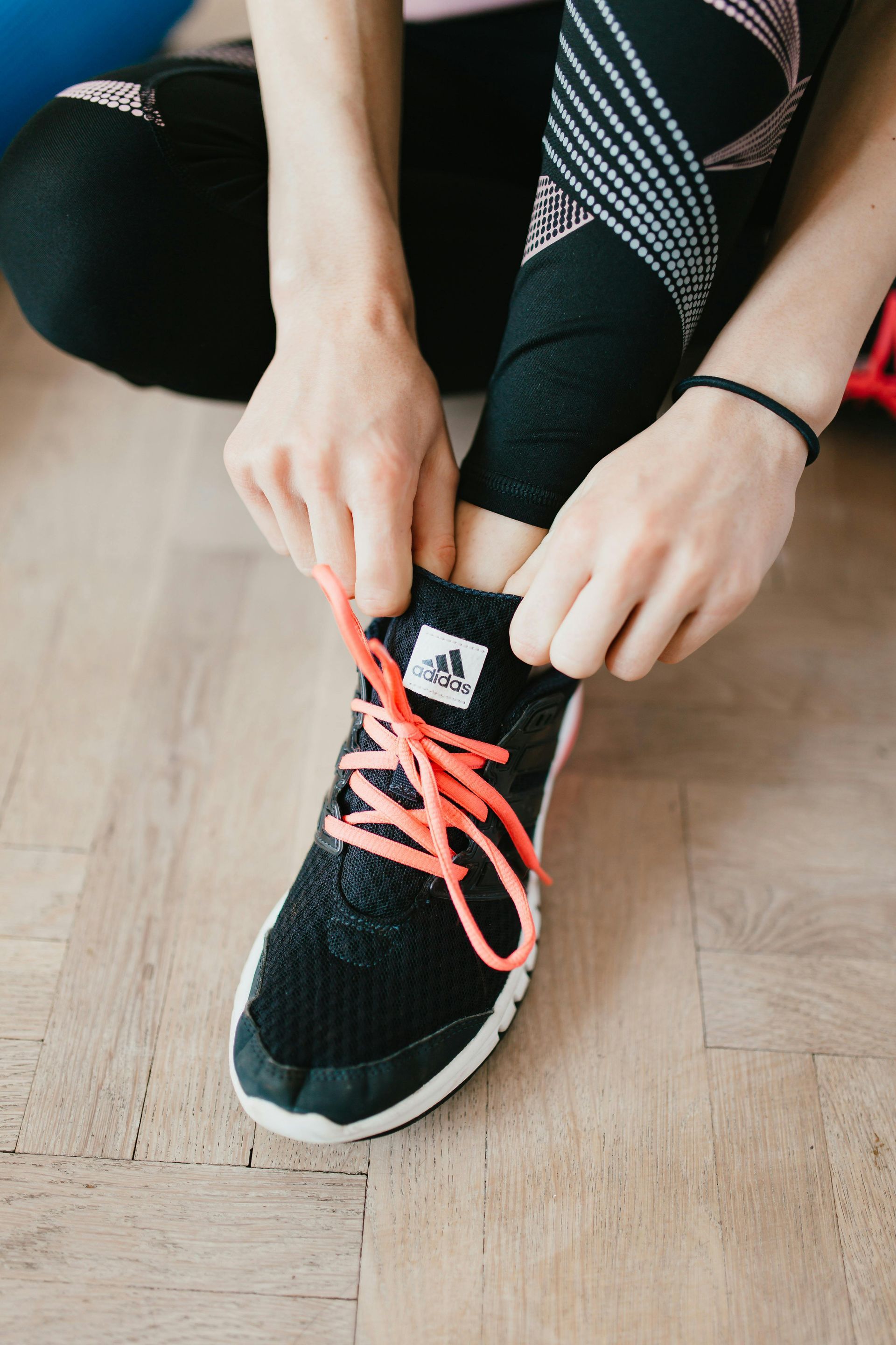 Hands tying bright coral shoelaces on a black sneaker on a wooden floor