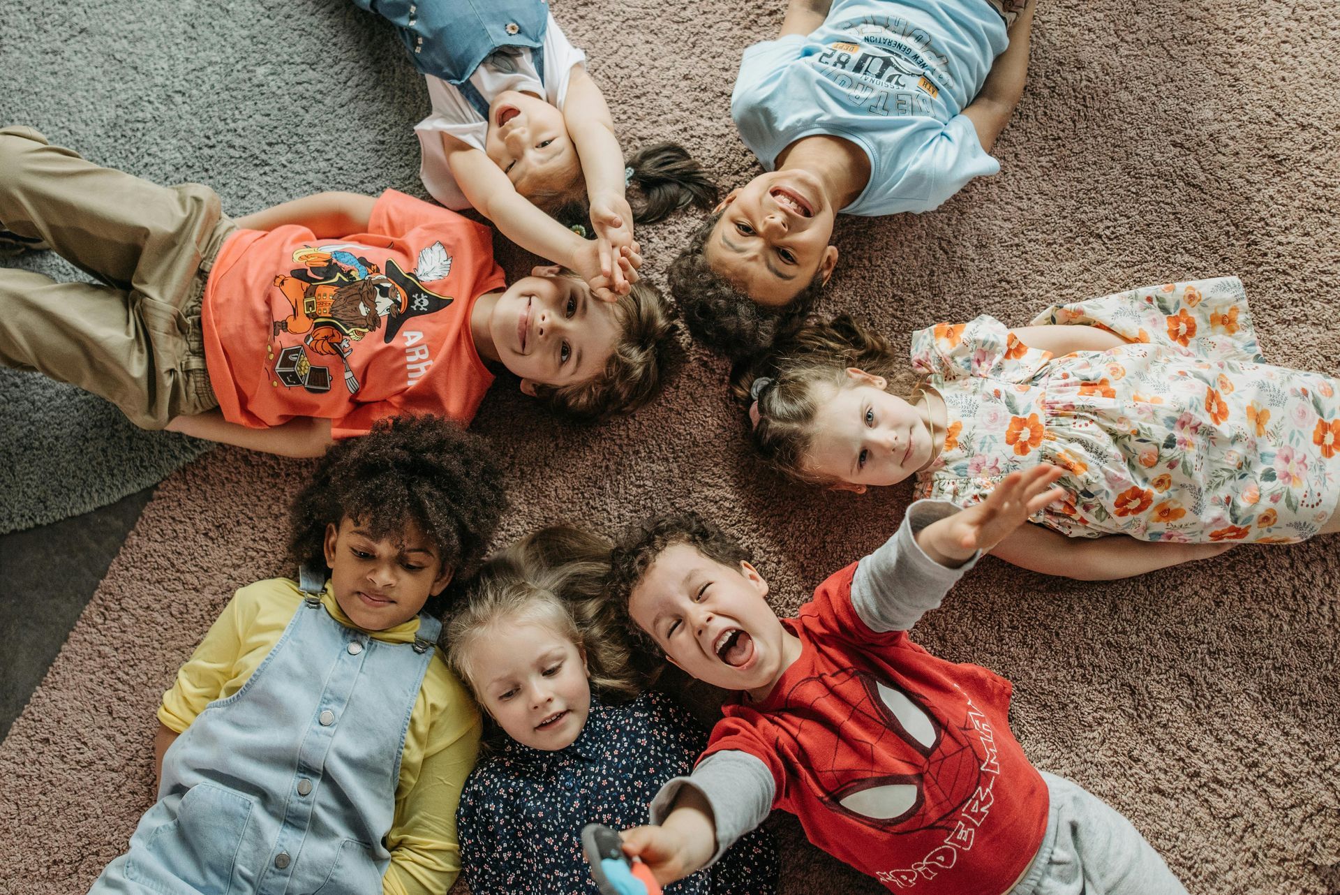 Five children lying in a circle on carpet, smiling up at the camera.