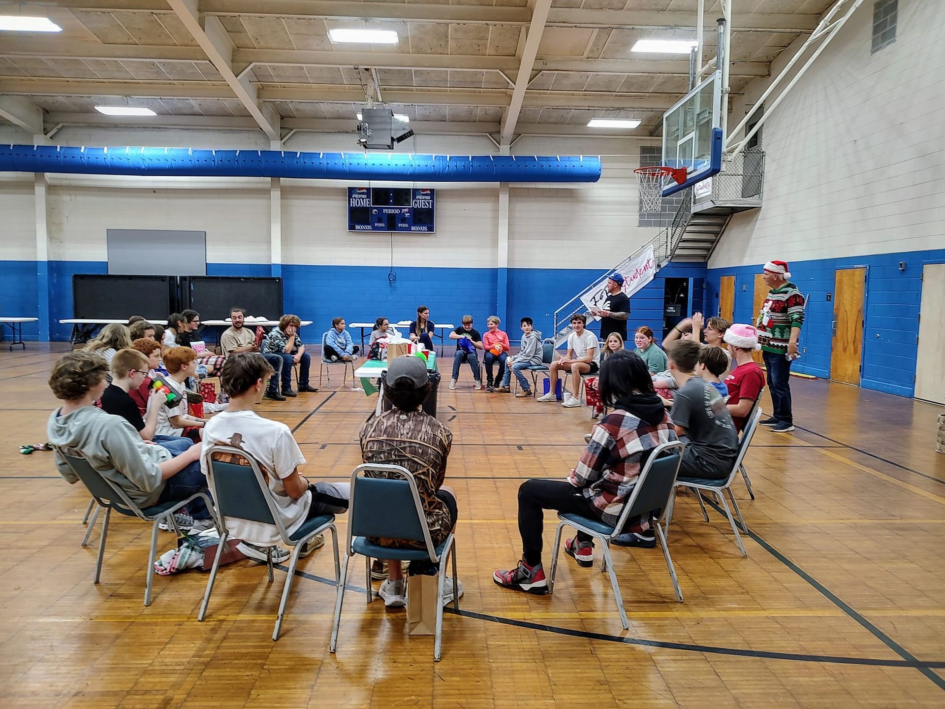 People seated in a gymnasium audience facing a stage area with blue walls and a basketball hoop.