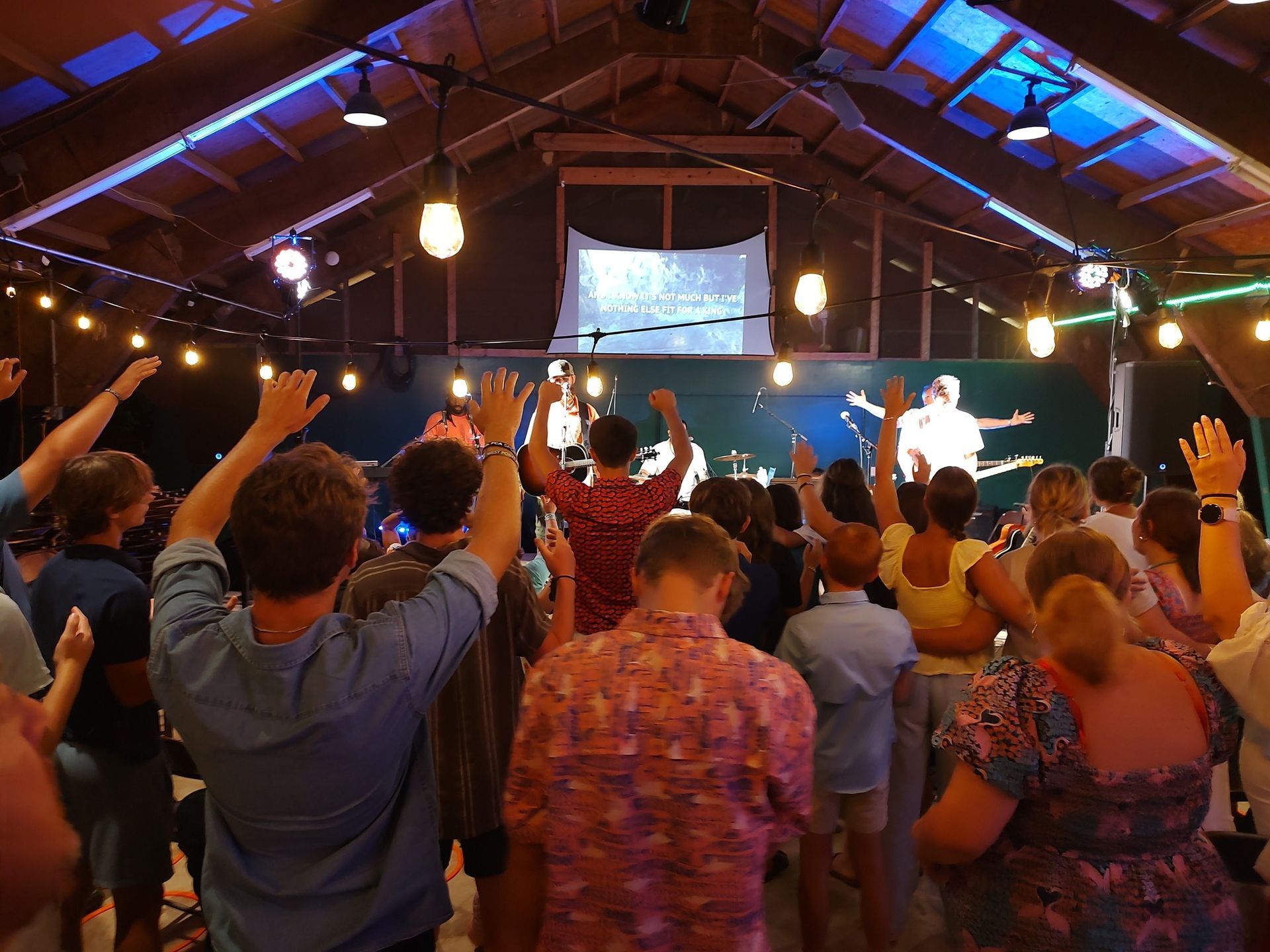 Crowd with raised hands at a concert under a lit wooden roof, facing a bright stage and screen