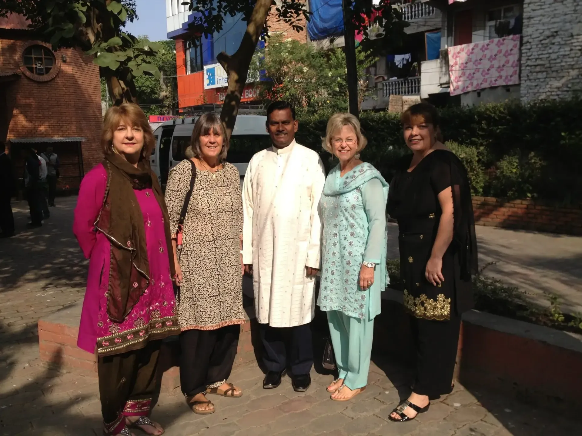 Five people standing outdoors in front of trees and buildings, posing in bright sunlight.