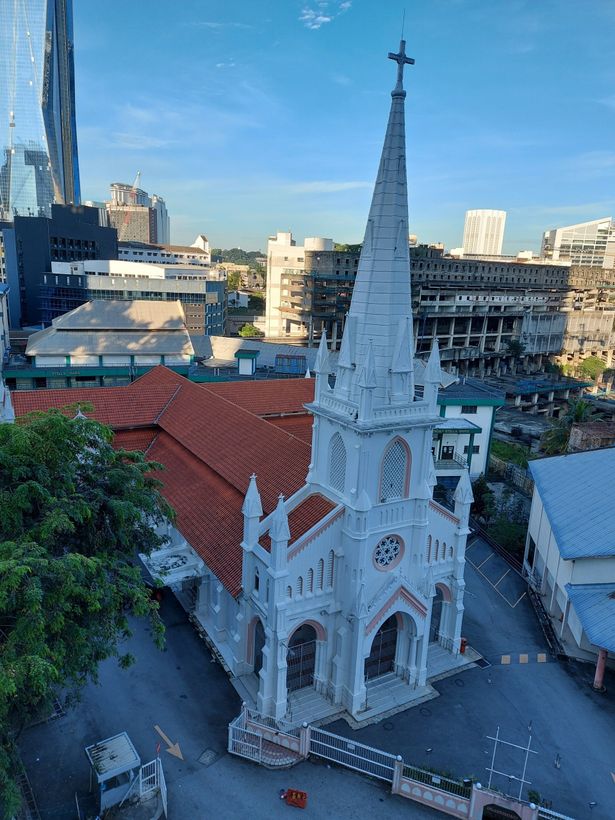 White church with tall spire, set in a city with red-tiled roof, surrounded by other buildings.