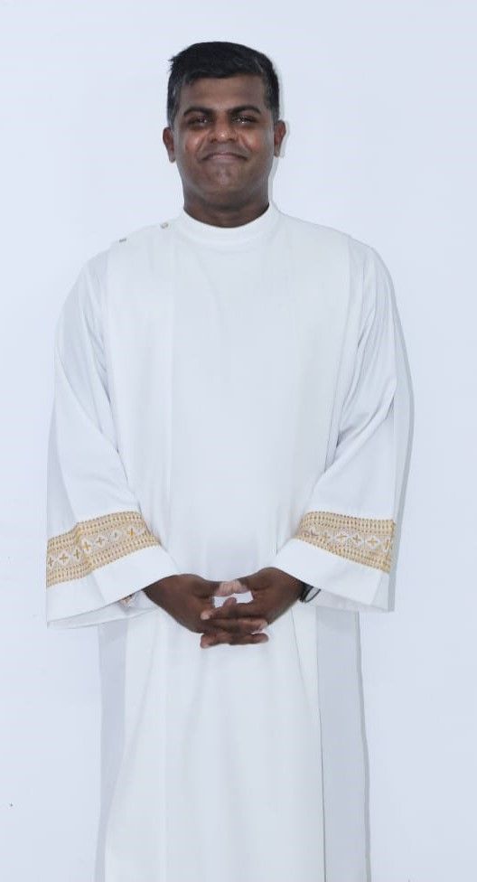 Man in white clerical robe with gold trim, hands clasped, smiling.