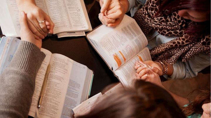 Hands clasped in prayer over open books. People in a group are holding hands and reading in a brightly lit room.