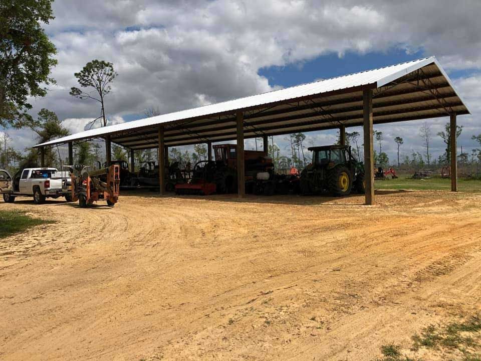 A row of tractors are parked under a covered area on a dirt road.