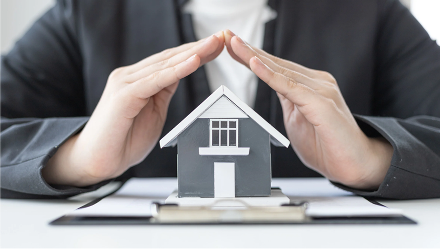 Hands sheltering a small house model on stacked books, symbolizing home protection and insurance.