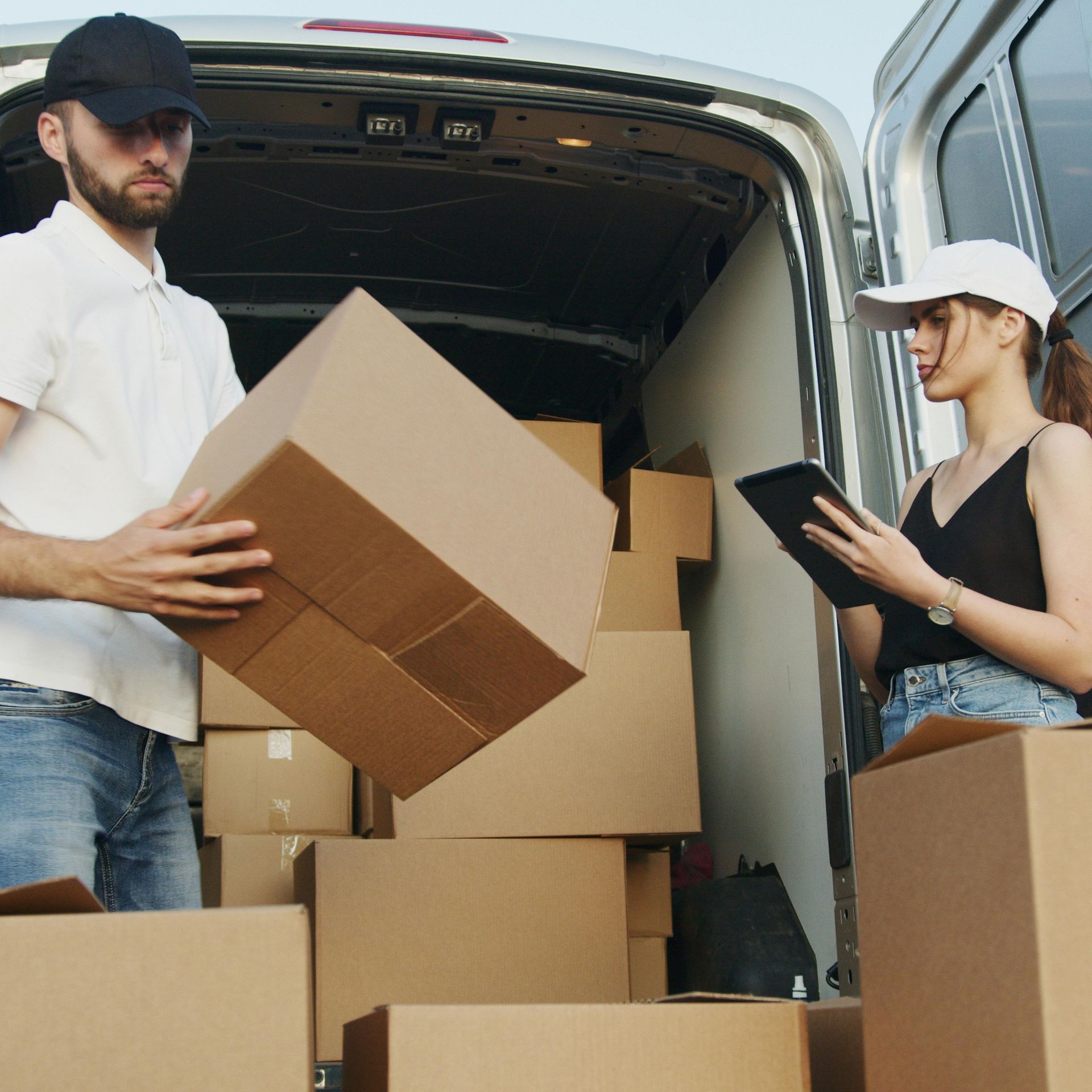 Two people behind a truck, the man is holding a cardbaord box while the woman is taking notes on a tablet.