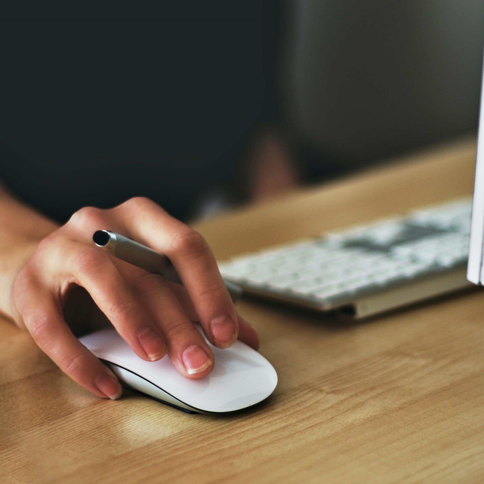 A hand on a computer mouse with a pen inbetween the fingers.