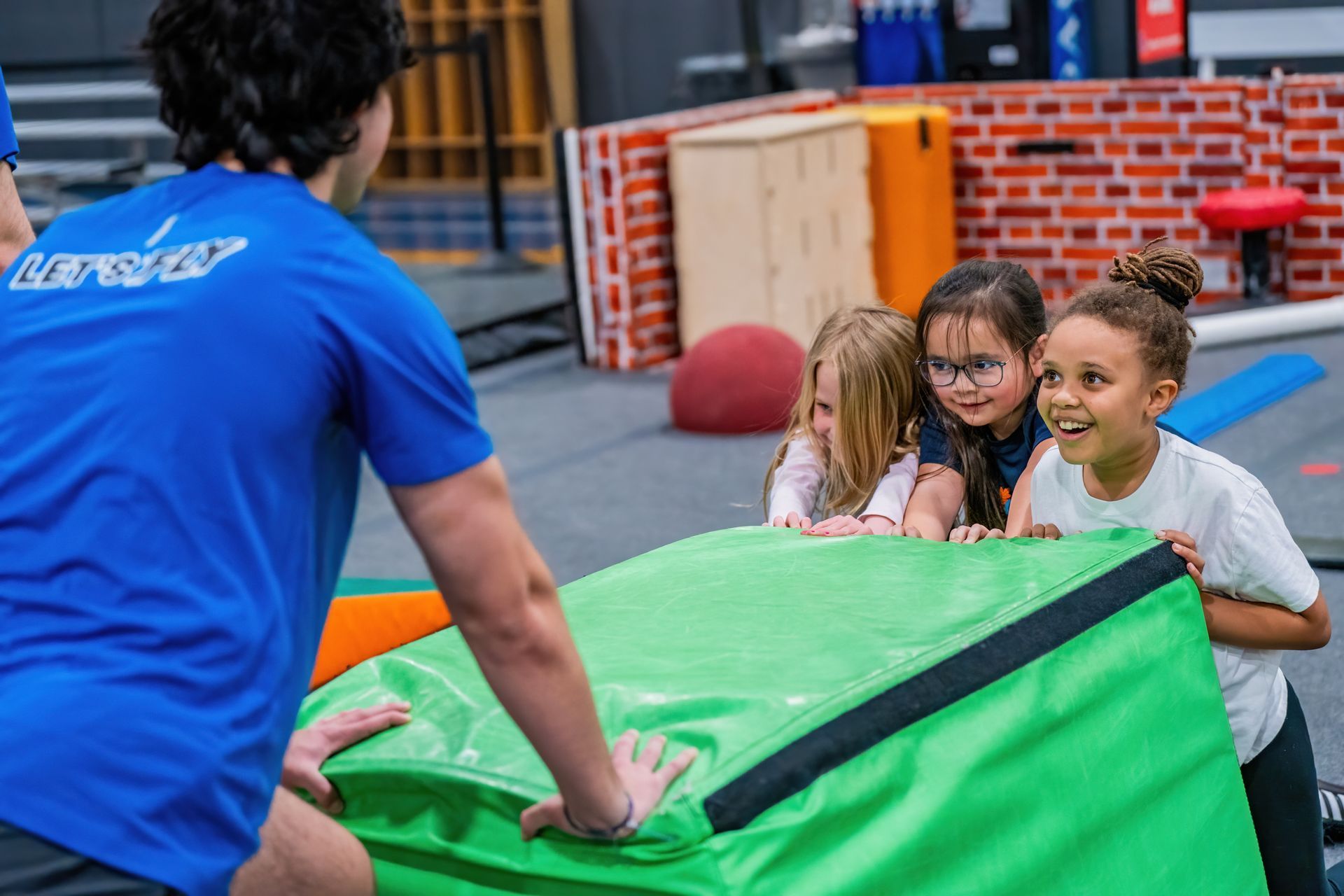 Instructor pushing green foam block as children reach and smile in an indoor play area