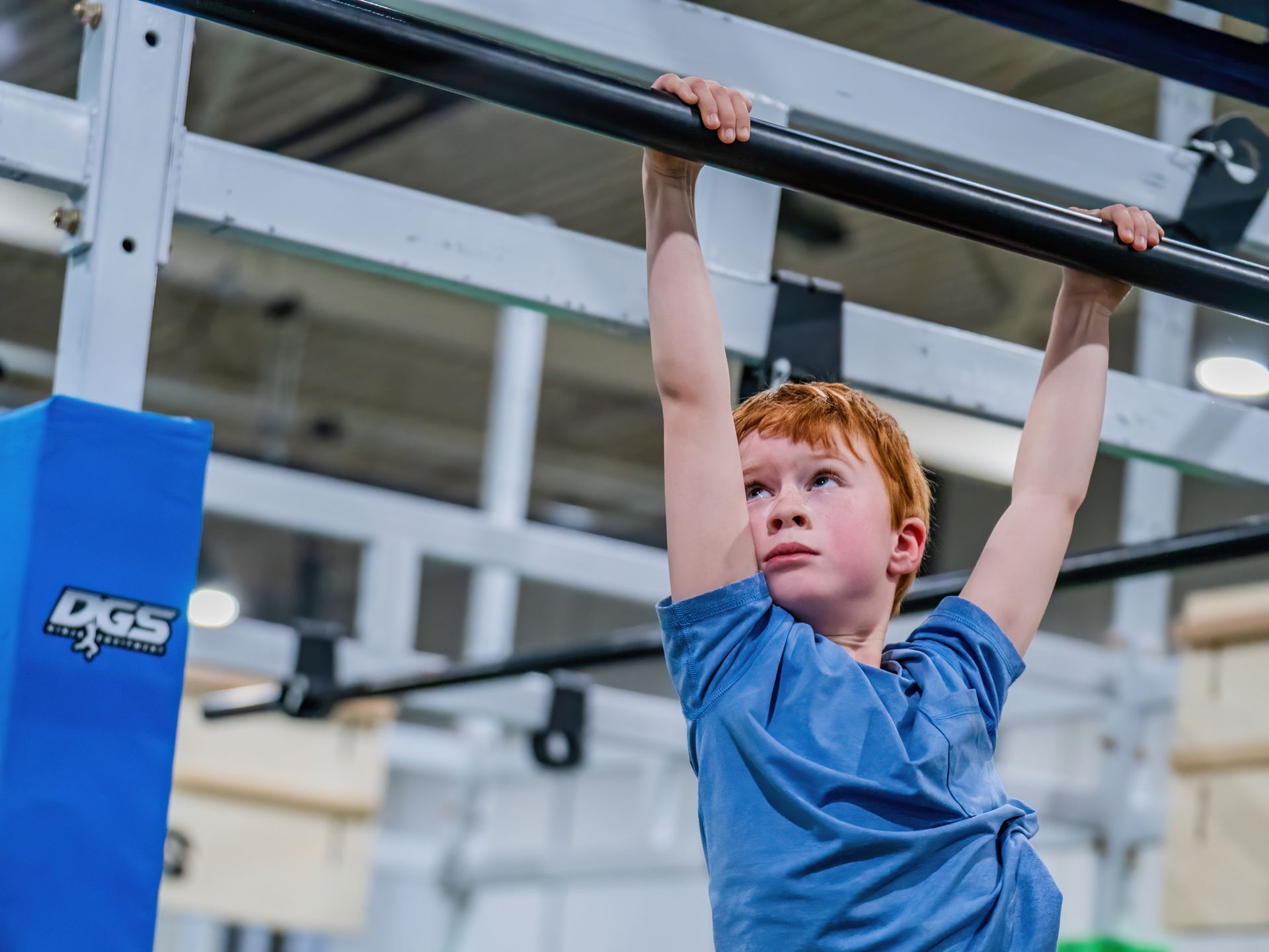 Child hanging from a monkey bar indoors, wearing a blue shirt and looking upward