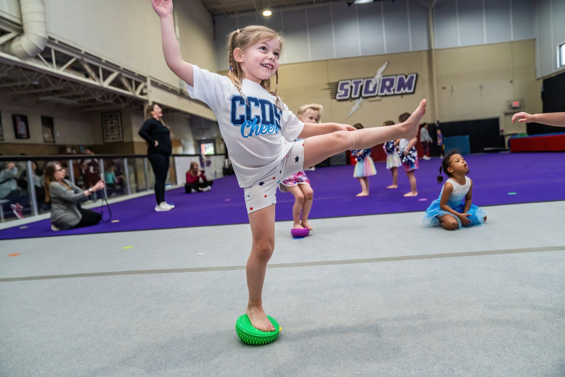 Child in a white leotard balances on one leg in a gymnasium, holding an outstretched arm with a green ball underfoot