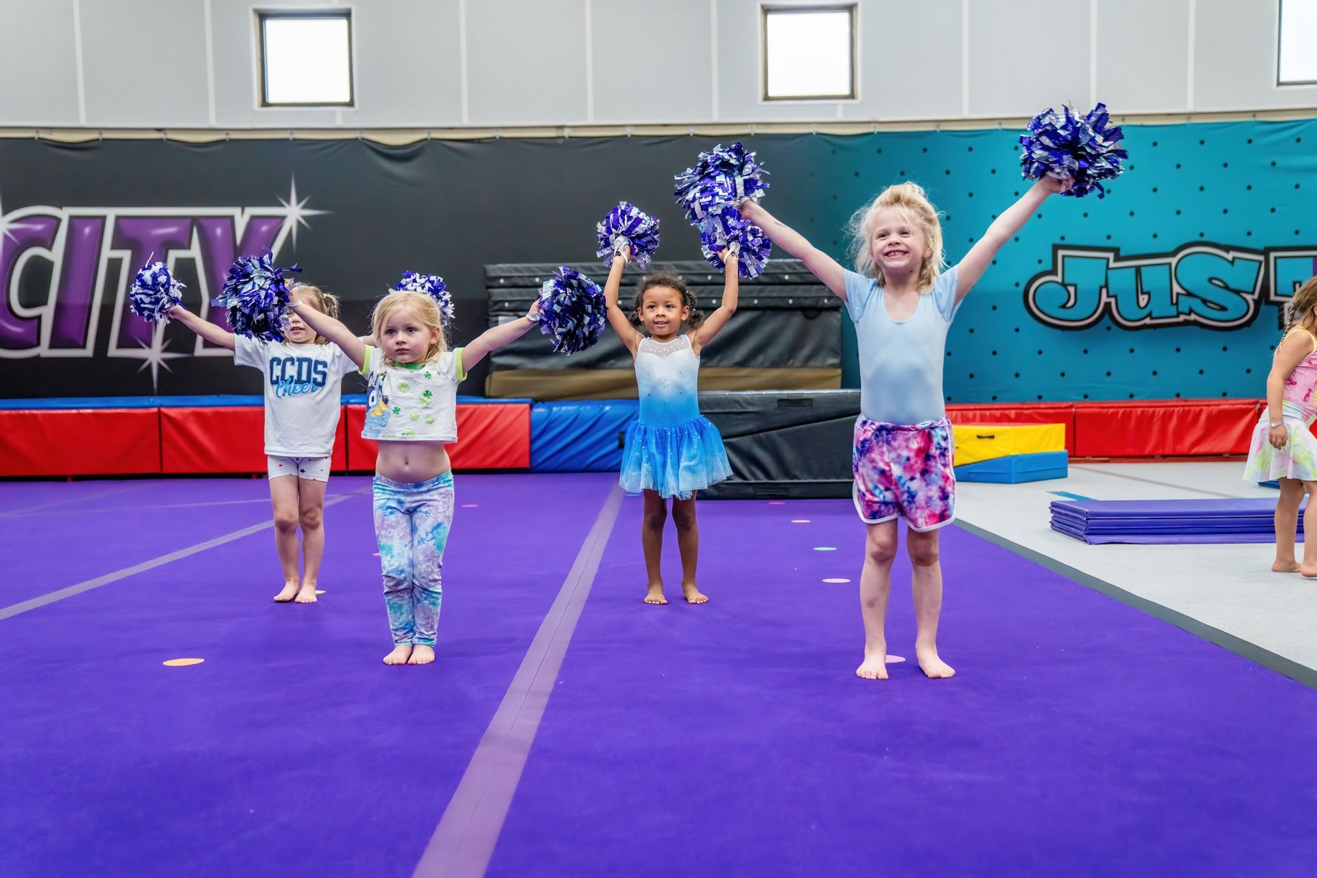 Four children cheering with pom-poms on a purple gymnastics mat in a gymnasium