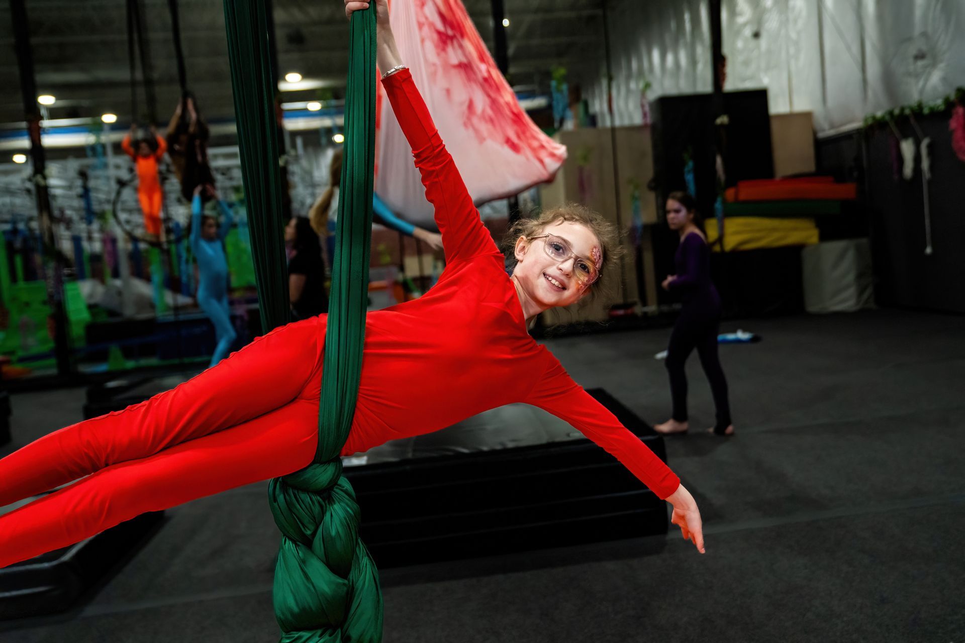 Person in a red outfit hanging horizontally from green aerial silks in an indoor gym.