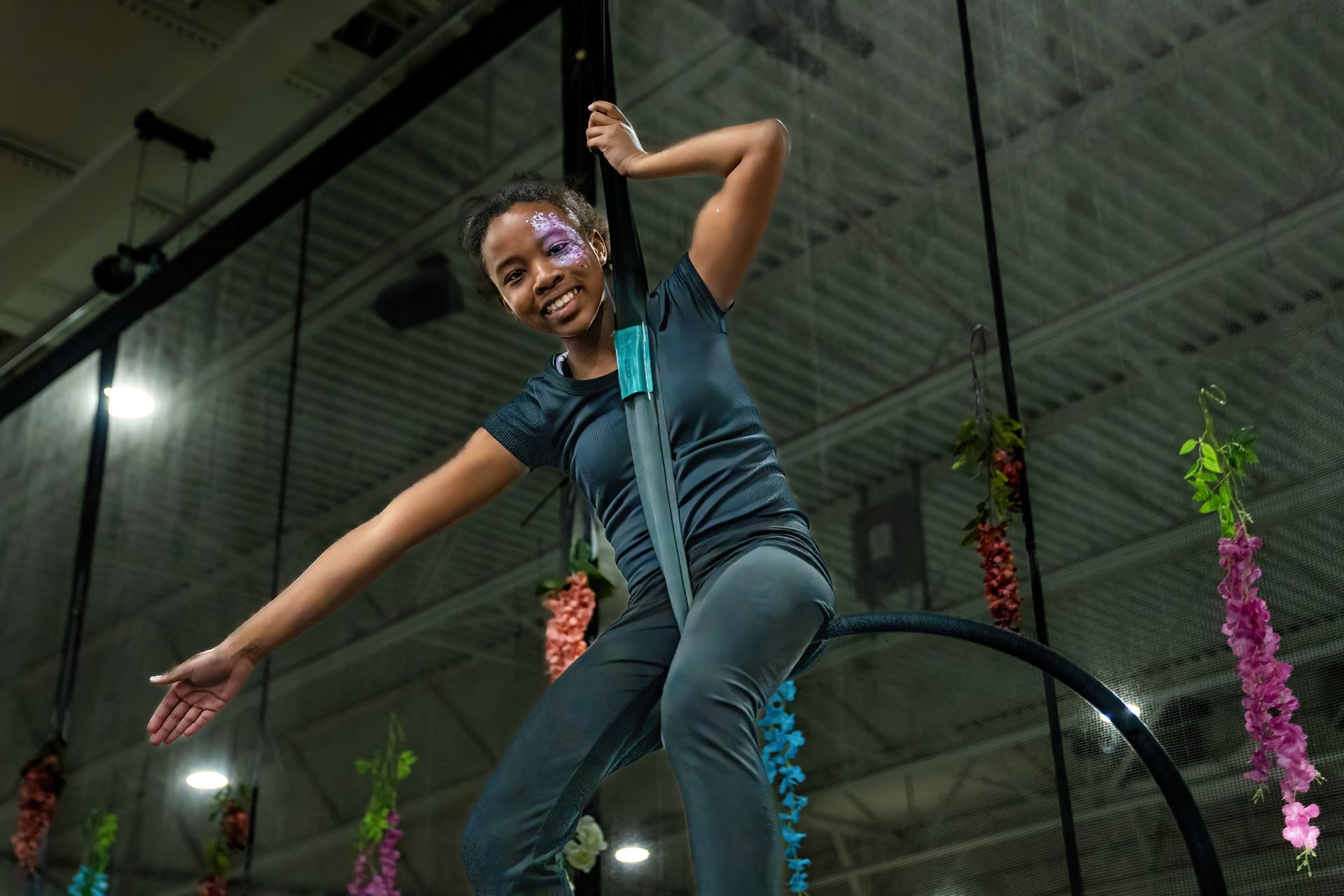 Smiling performer in teal outfit posing on indoor aerial hoop in a gym-like space