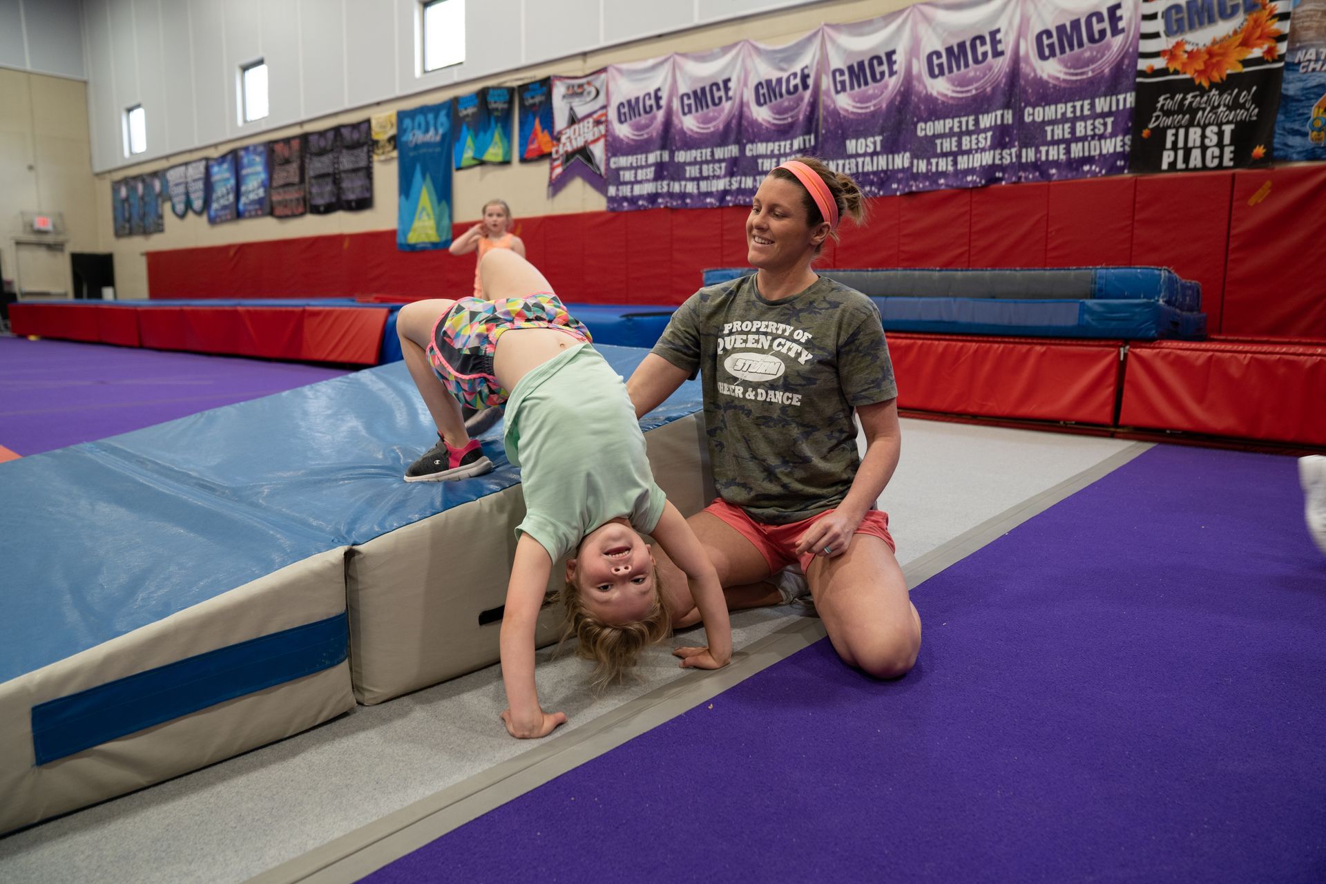 Gymnastics coach assists a child doing a backbend on a purple mat in a gymnasium
