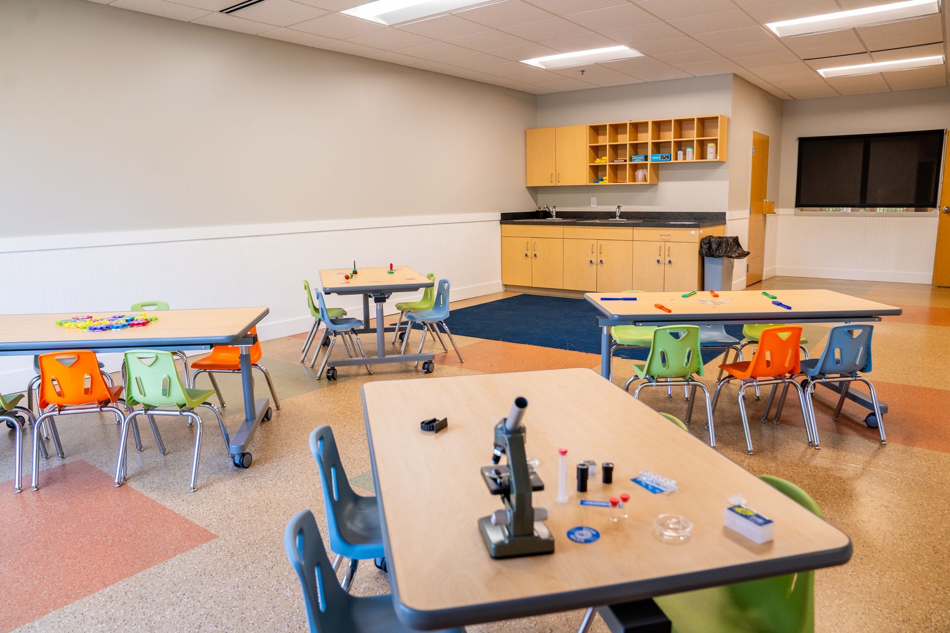 A brightly lit classroom with desks and stools, a water fountain, cabinets, and a wall that says 