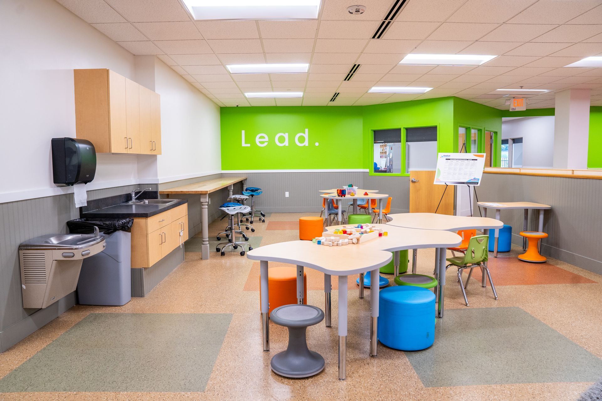 Bright classroom with green walls, tables, stools, cabinets, and a “Lead.” sign on the back wall.