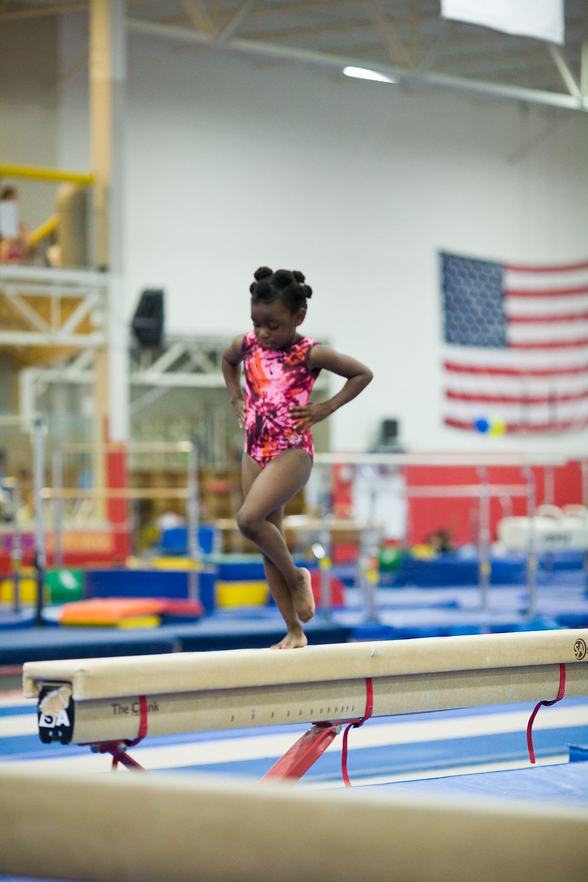 Gymnast balancing on a beam in a gym, with an American flag in the background.