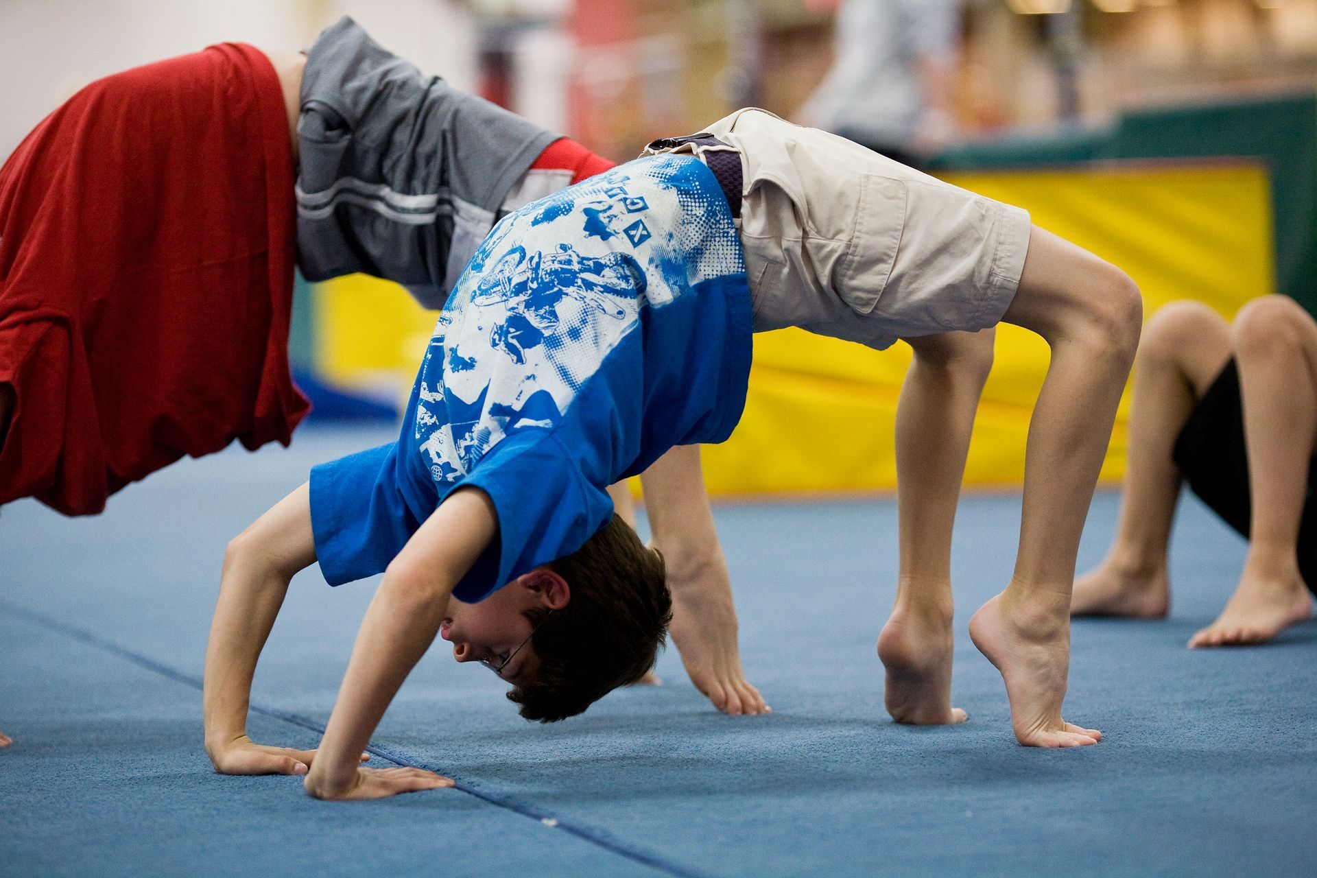 Children doing backbends on a blue mat in a gym class, wearing colorful shirts and shorts