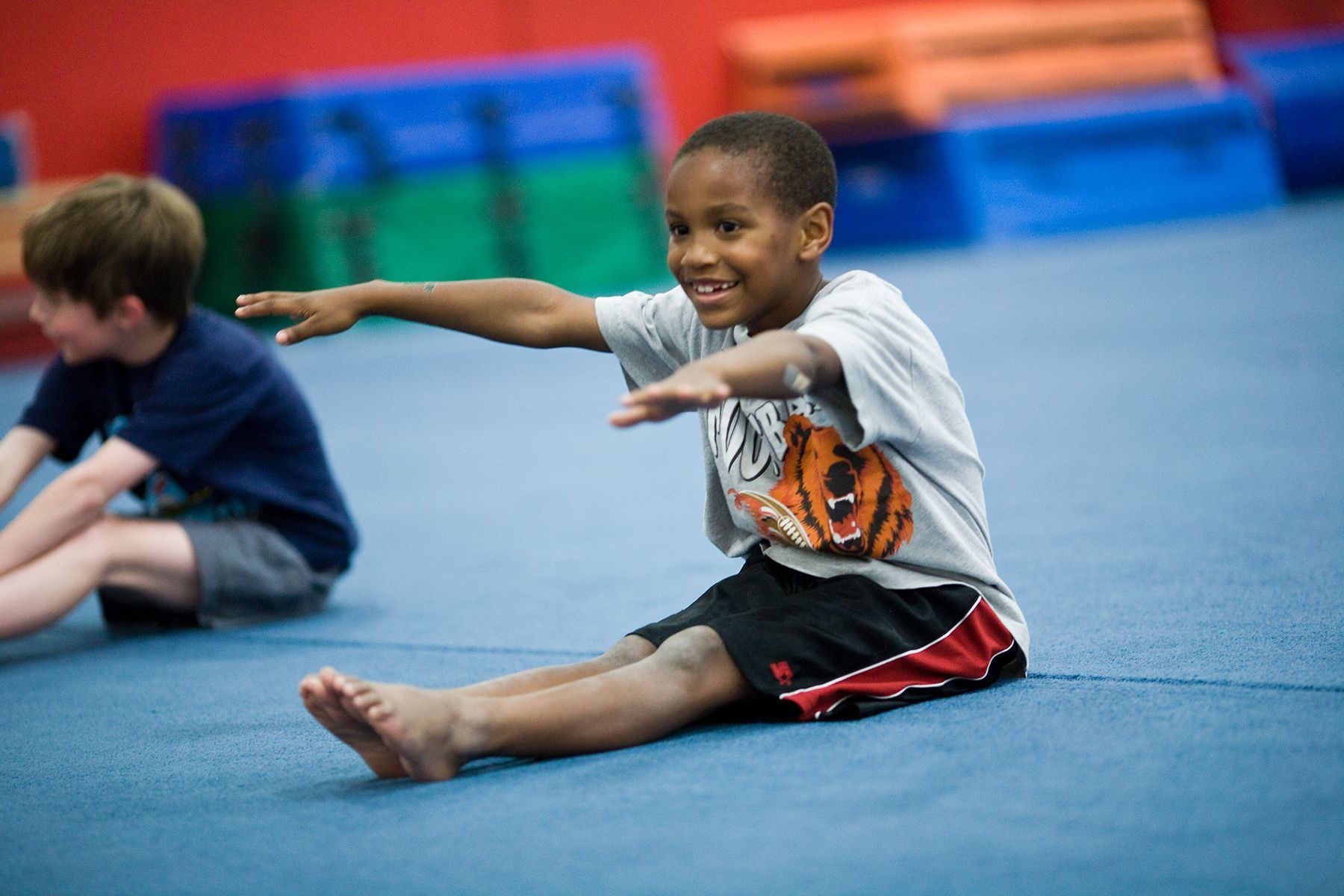 Boy sitting on gym mat, smiling and stretching arms forward during a class