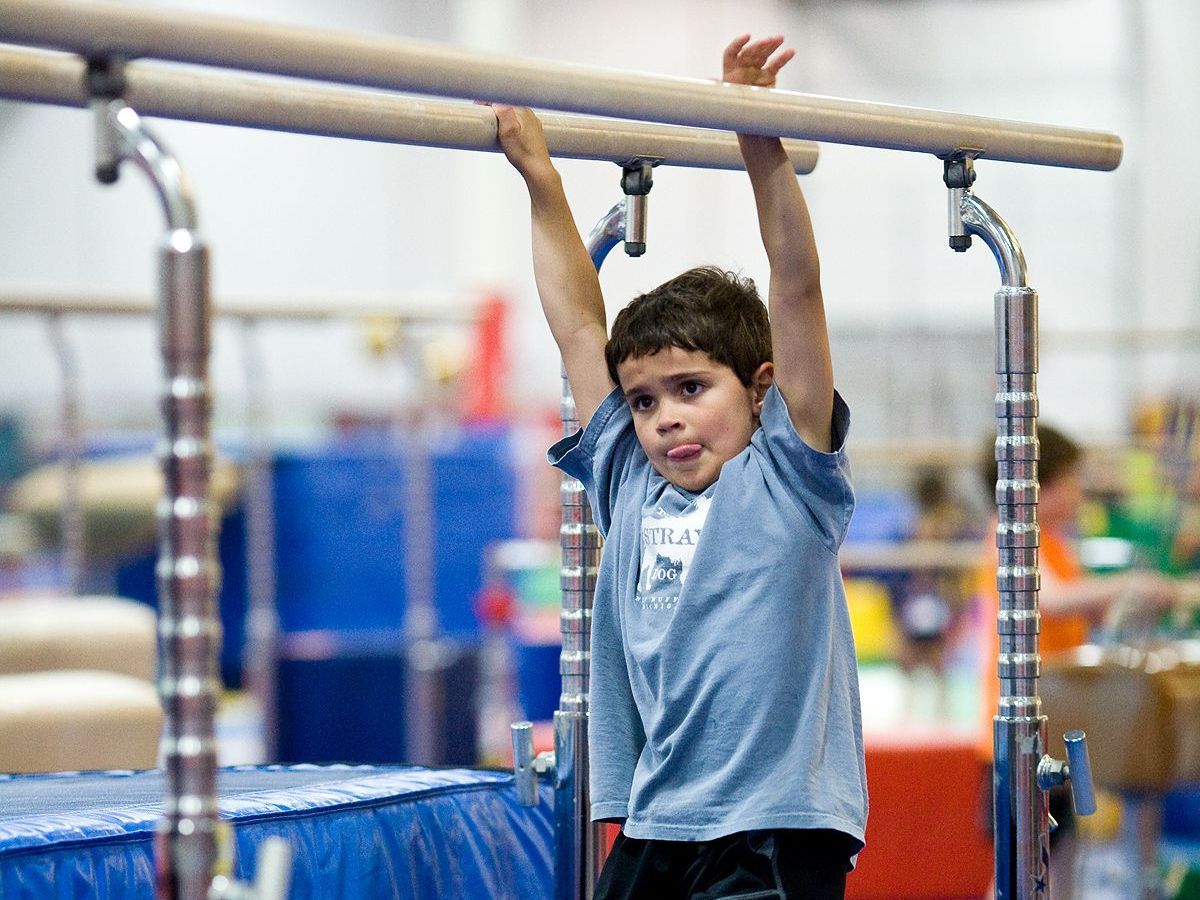 Child hanging from a gymnastics bar in a gym, with blue mats and equipment in the background