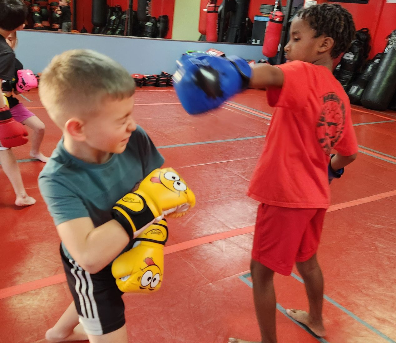 Two children sparring with boxing gloves in a gym, one punching while the other blocks.