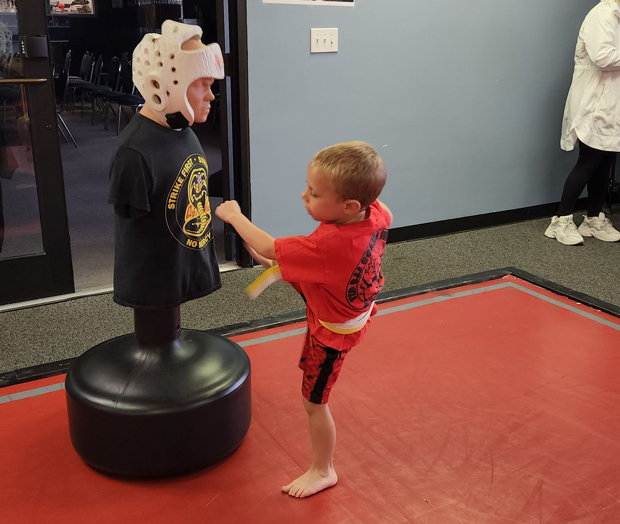 Child practicing kicks on a martial arts training dummy in a gym.