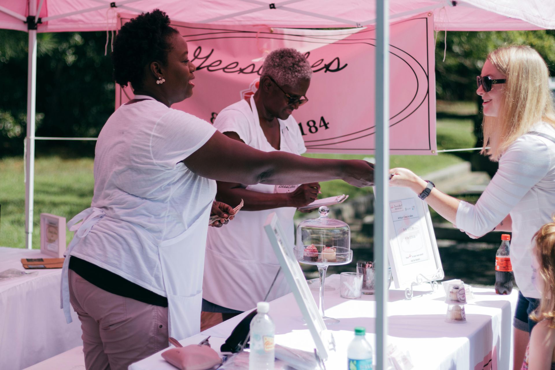 Two women exchange items across a booth at an outdoor pink vendor tent.