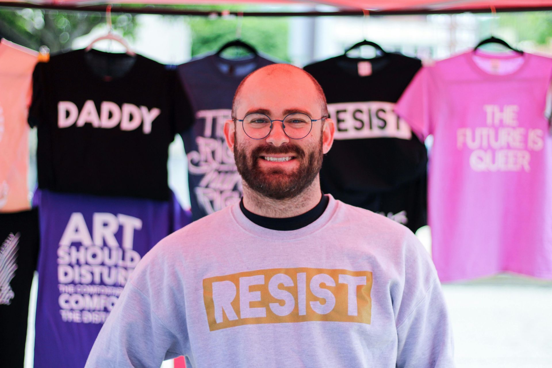 Smiling person in a gray RESIST sweatshirt standing before protest slogan shirts at a booth