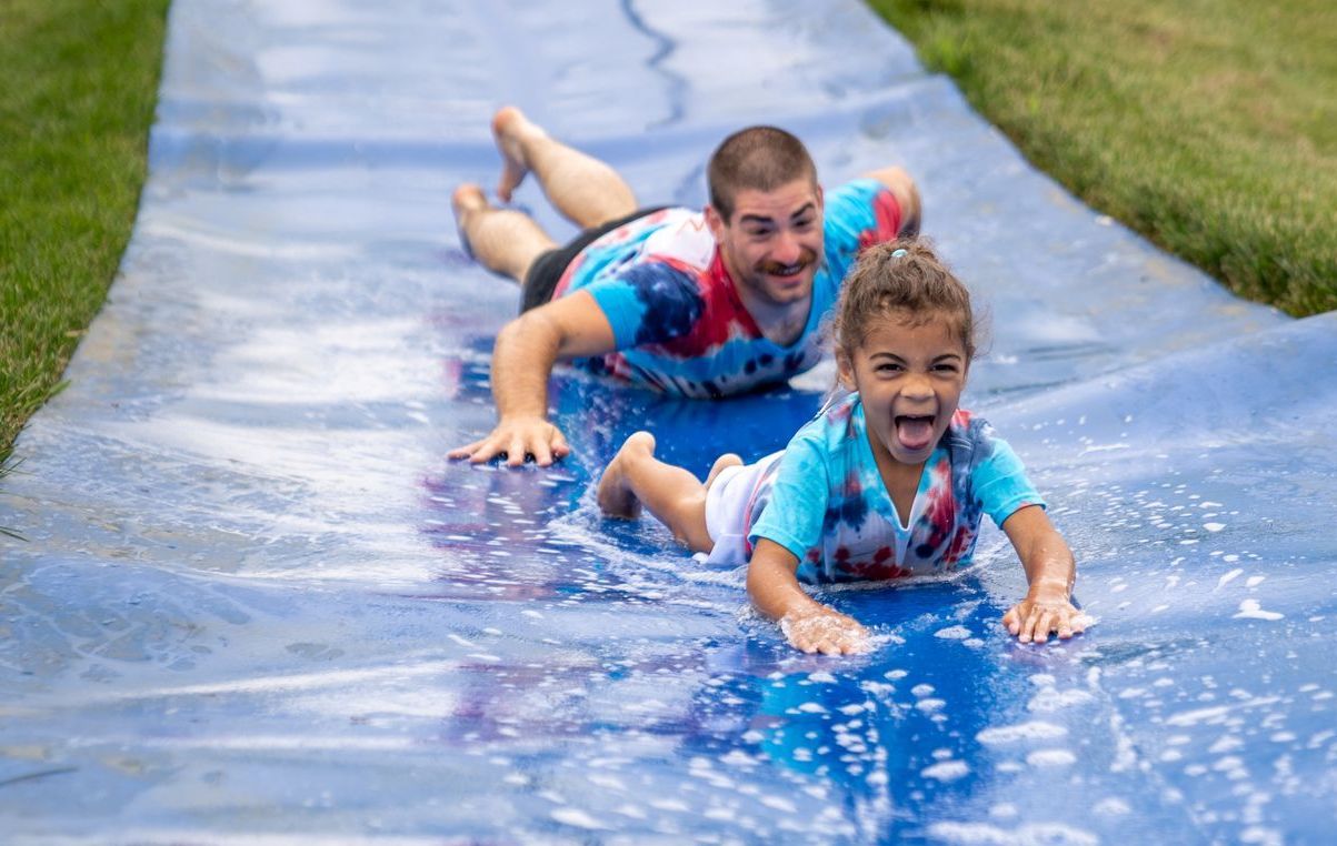 Two children sliding down a blue water slide, splashing and smiling outdoors