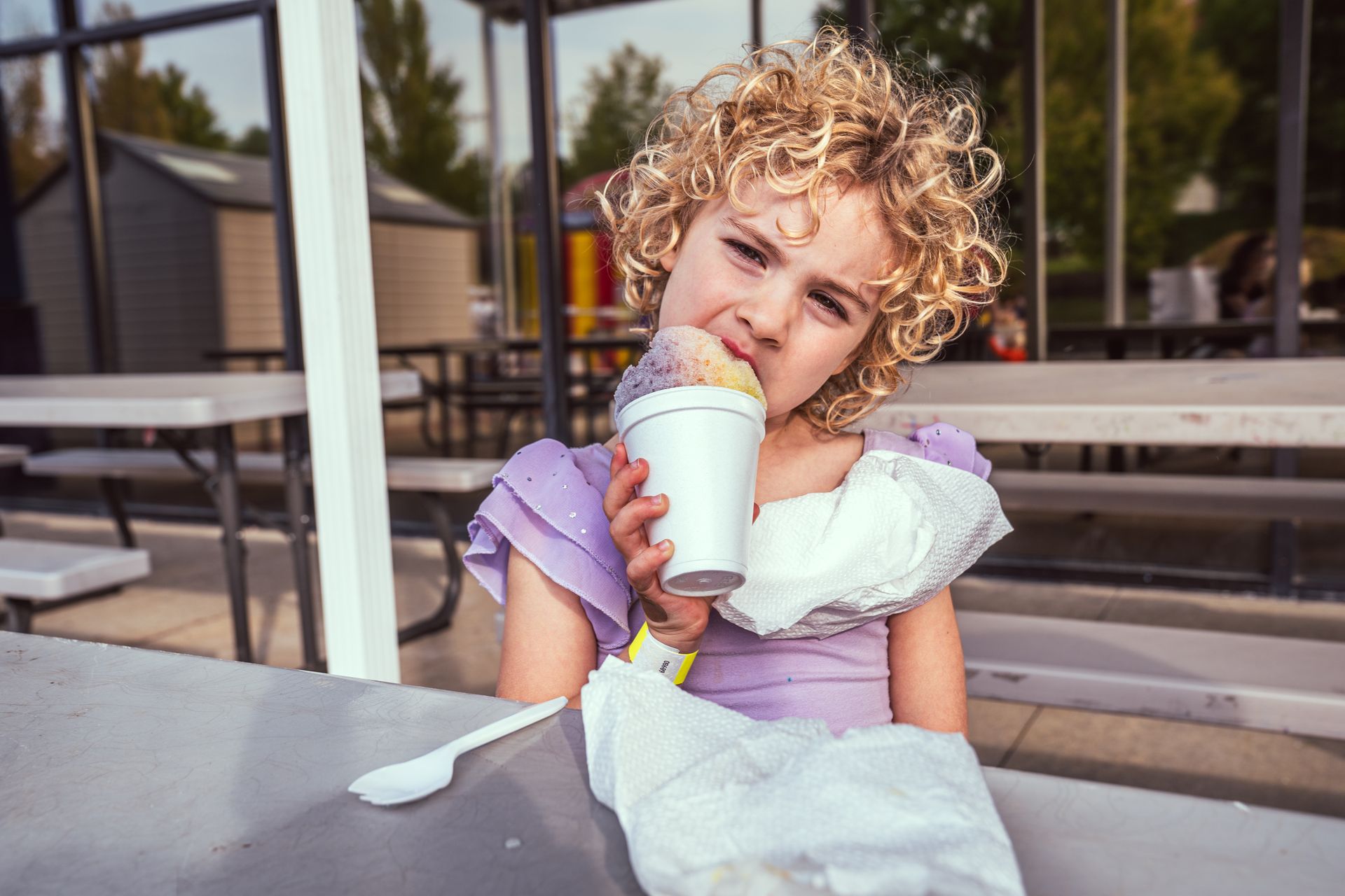Smiling child in a purple shirt drinking from a white cup at an outdoor café table