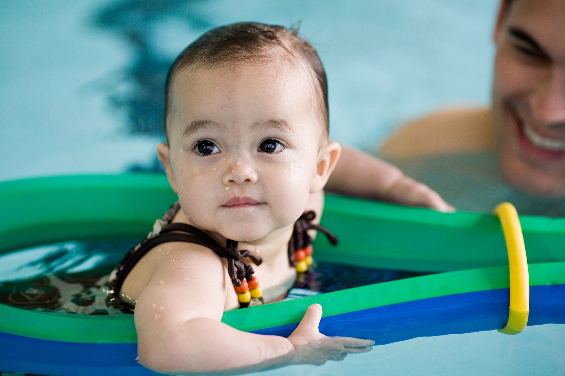 Baby in a green float in a swimming pool, reaching forward with a calm expression.