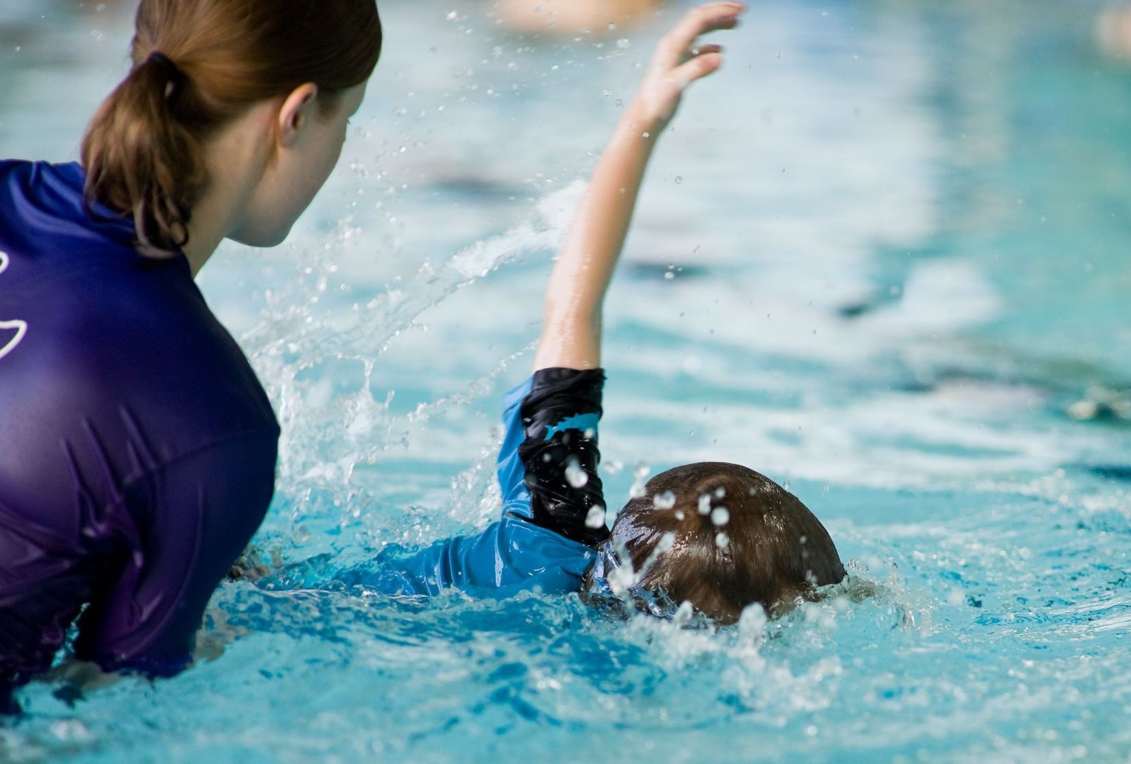 Swimmer in blue splashing in a pool while a coach watches nearby