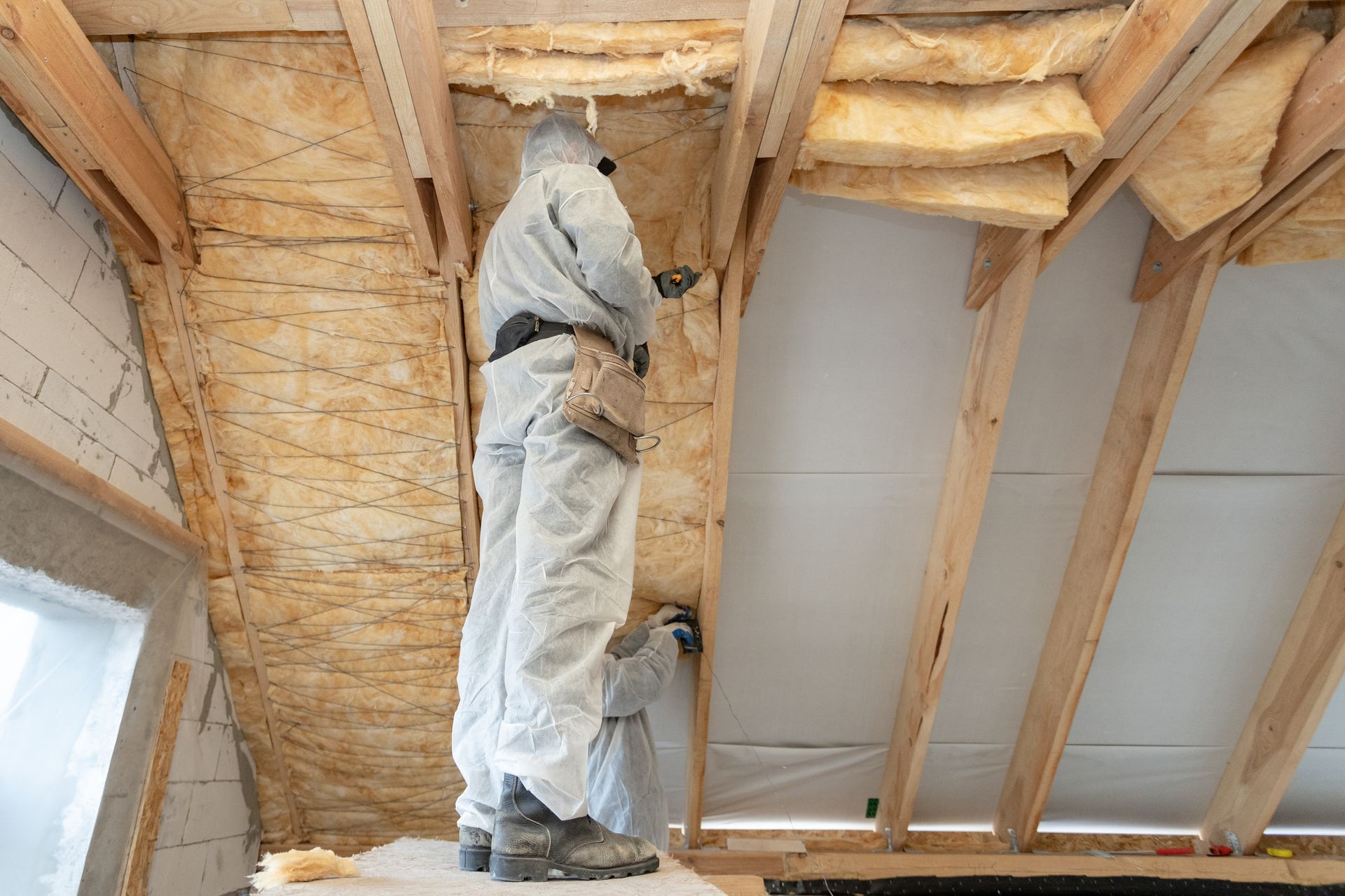 Person in white protective suit spraying insulation into an attic ceiling cavity.