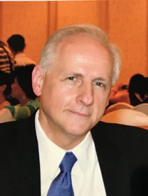 Portrait of a man in a dark suit and blue tie, seated against a blurred warm-toned background.