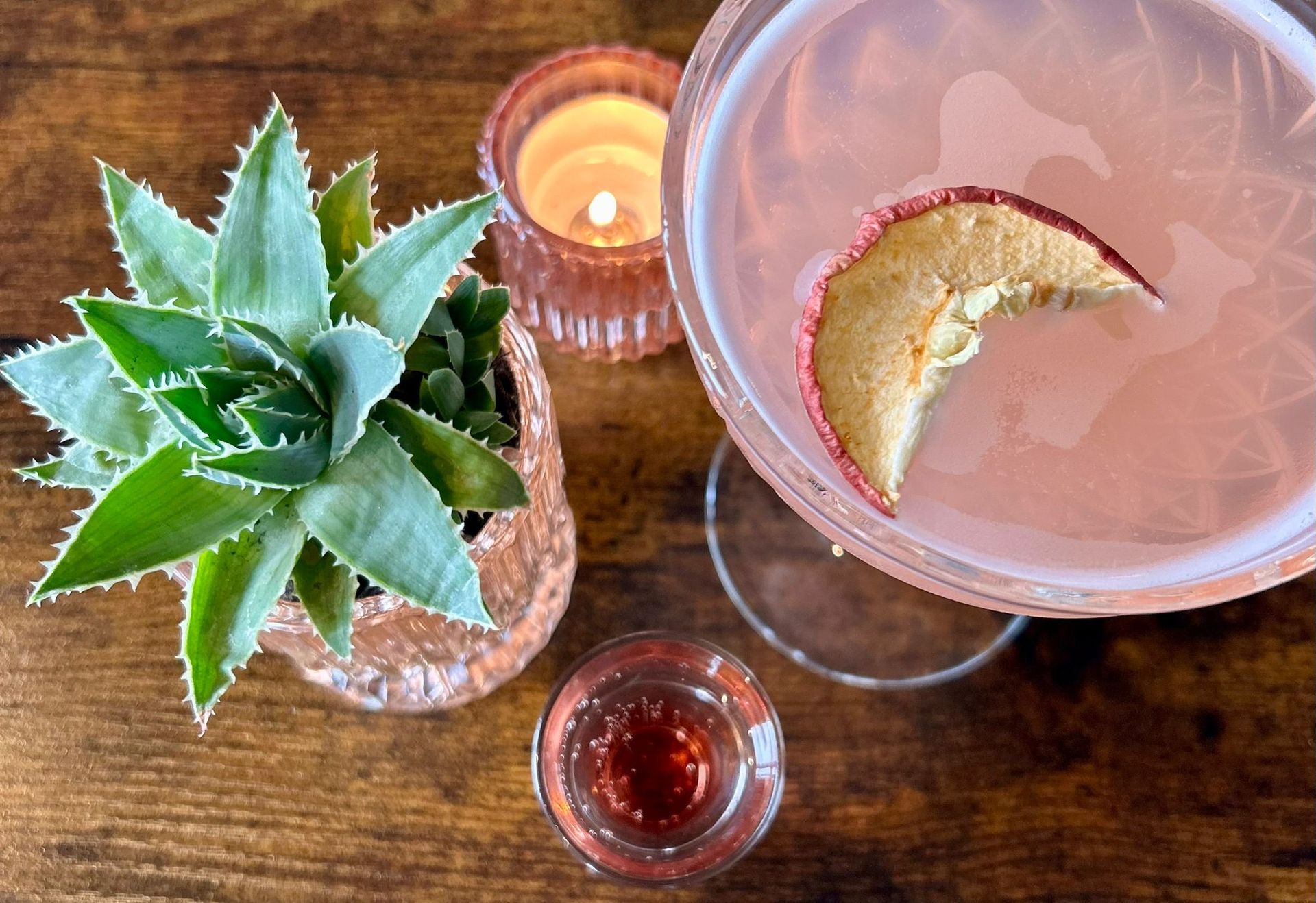 A cocktail with a dried fruit garnish next to a small succulent and a lit candle on a wooden table.