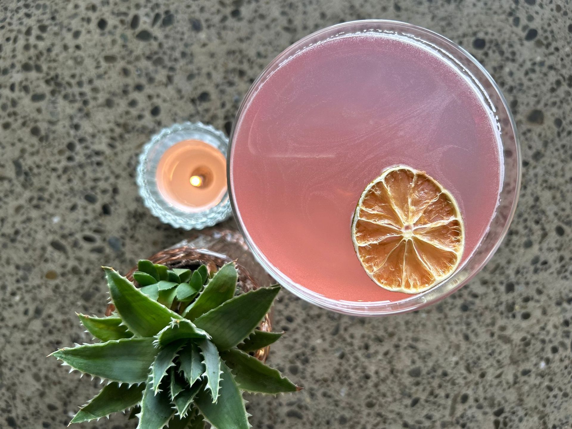 A pink cocktail garnished with a dried lime slice next to a small succulent and a votive candle on a gray stone surface.