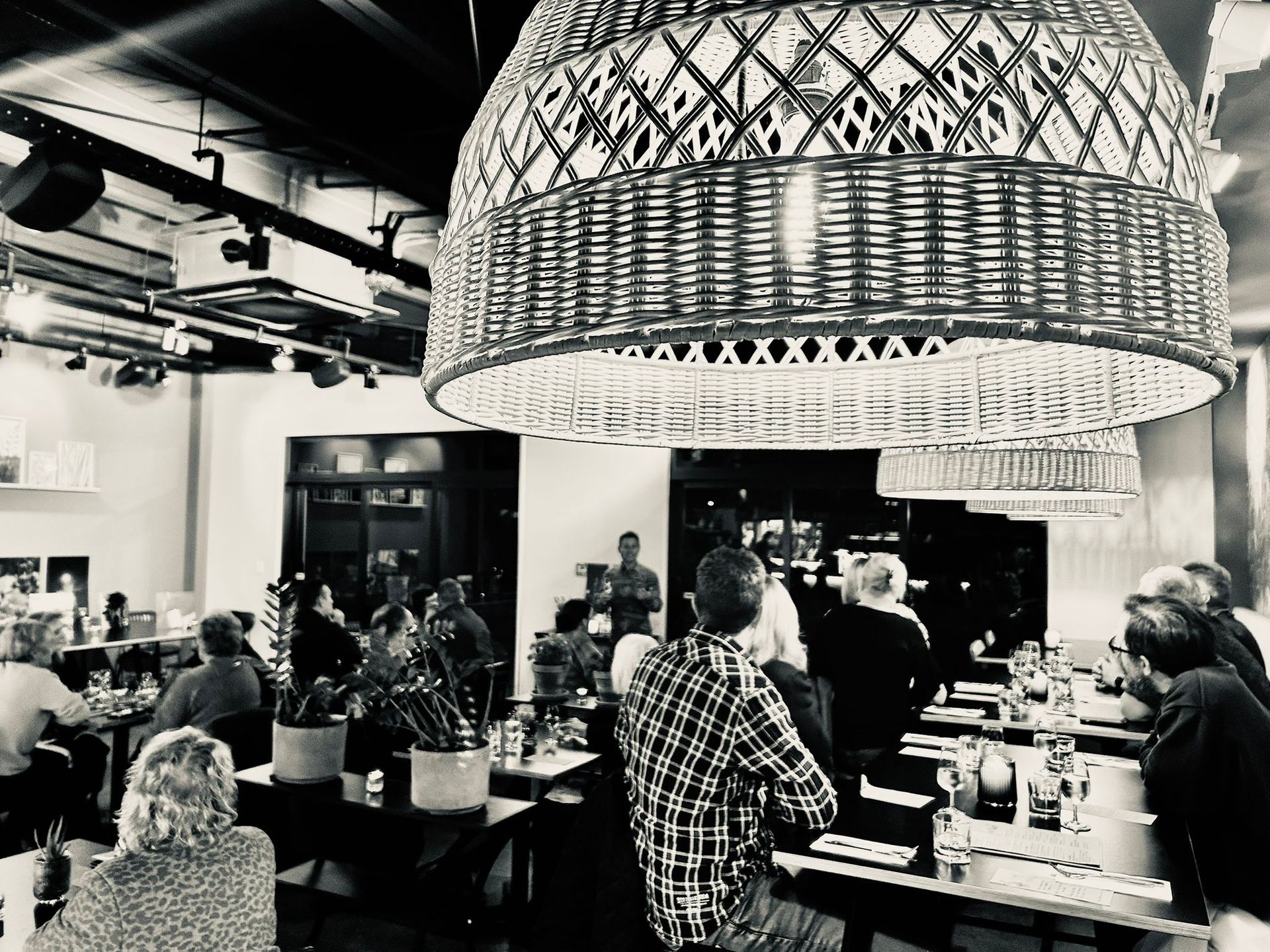 A black and white view of a dimly lit restaurant featuring a prominent, large woven pendant light above seated diners.