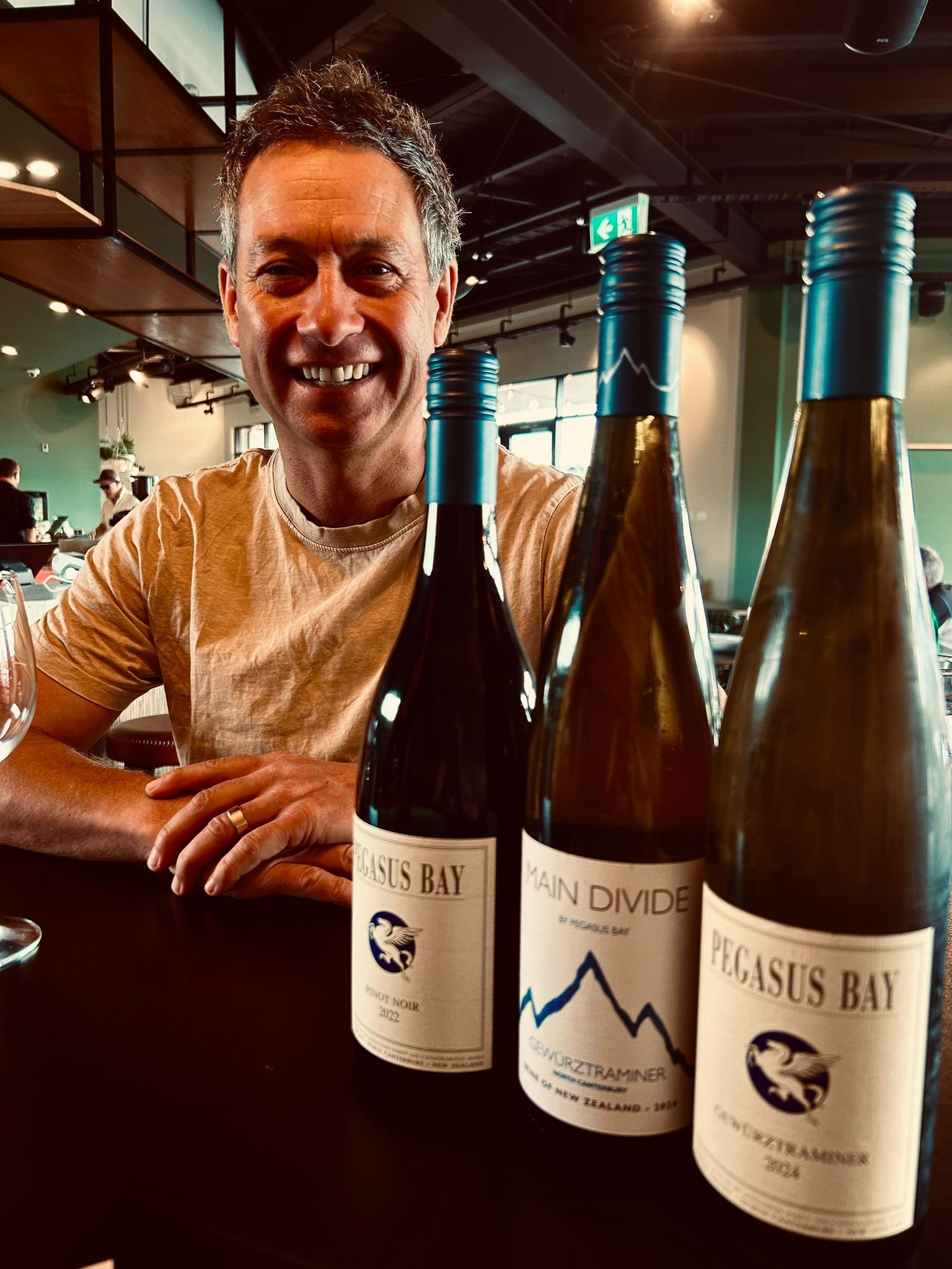 A smiling person sits at a table with three bottles of Pegasus Bay wine in a brightly lit indoor setting.