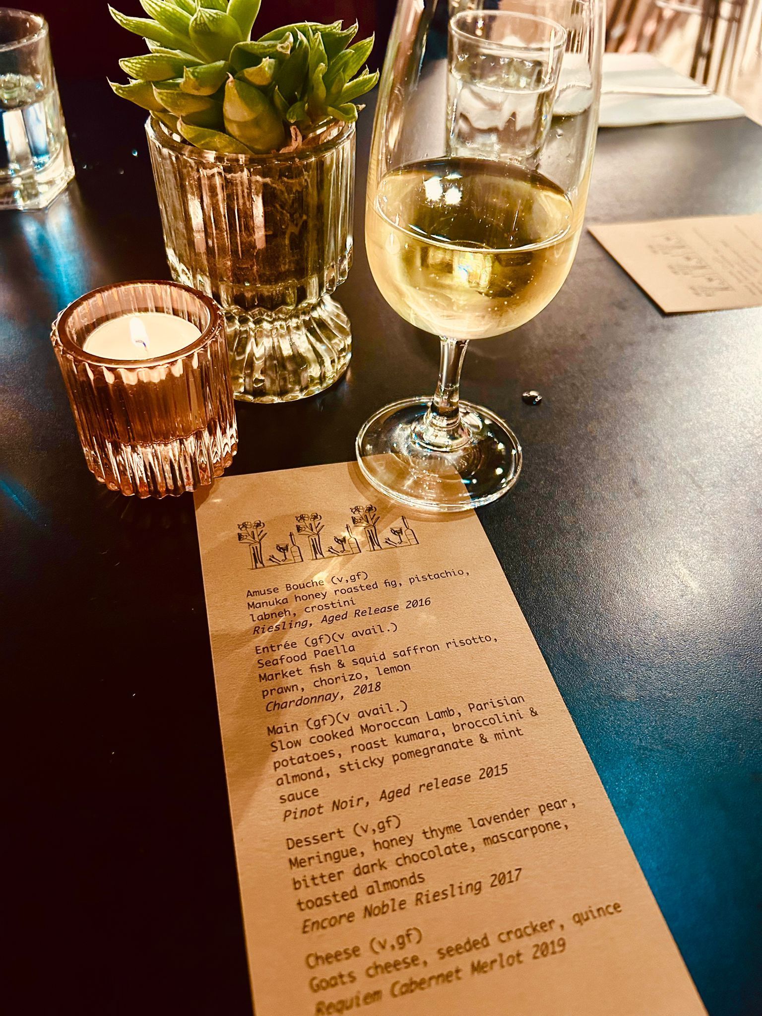 A close-up of a dimly lit restaurant table with a menu, a glass of white wine, a lit candle, and a small potted succulent.